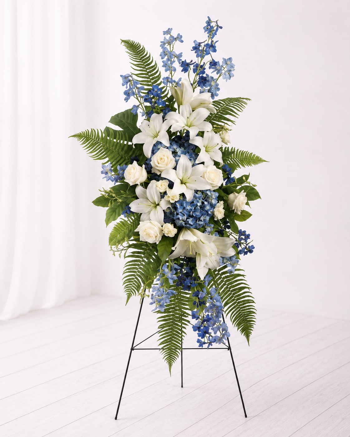 Blue Skies Standing Spray - Elegant and peaceful, the Blue Skies Standing Sympathy Spray is a graceful tribute designed to honor and remember a loved one. Featuring stunning white lilies, soft white roses, blue hydrangea, delicate blue delphinium, and lush cascading greenery, this standing arrangement offers a comforting expression of love, peace, and heartfelt sympathy. Displayed on an easel, it creates a beautiful and meaningful memorial presentation for funeral services or celebrations of life.