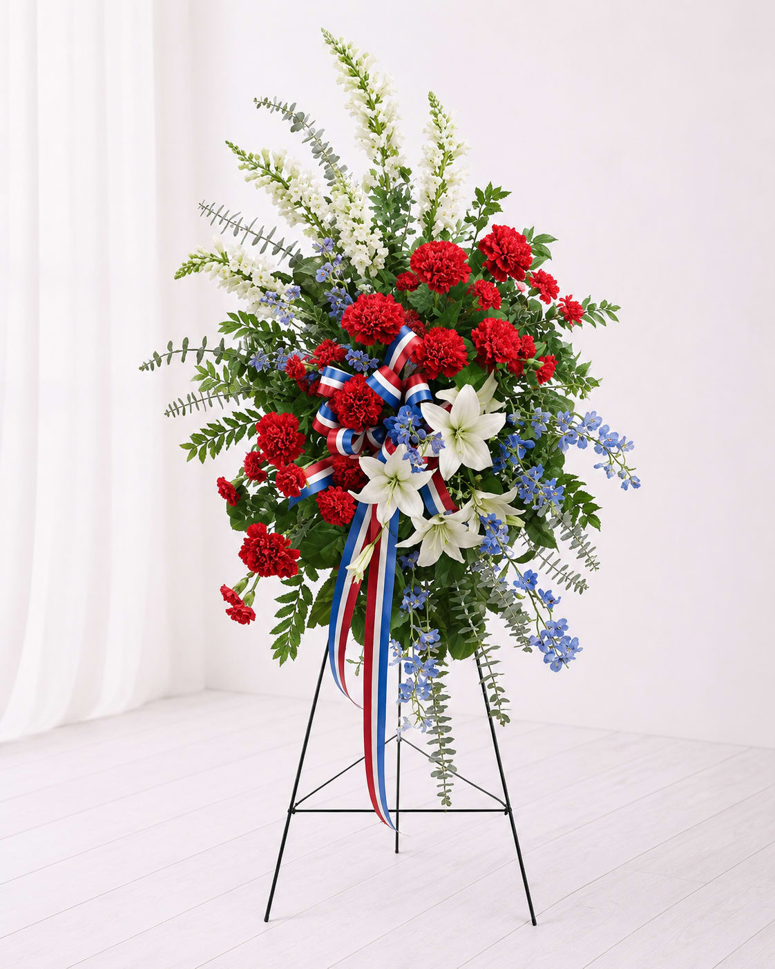 Patriotic Honor Standing Spray - Honor a life of courage, service, and love with the Patriotic Honor Standing Spray. This striking red, white, and blue tribute features vibrant red carnations, elegant white lilies and snapdragons, delicate blue delphinium, lush greenery, and a patriotic ribbon accent beautifully displayed on a standing easel. Designed to express deep respect, remembrance, and heartfelt sympathy, this arrangement is a meaningful tribute for veterans, first responders, or anyone proudly remembered with honor.