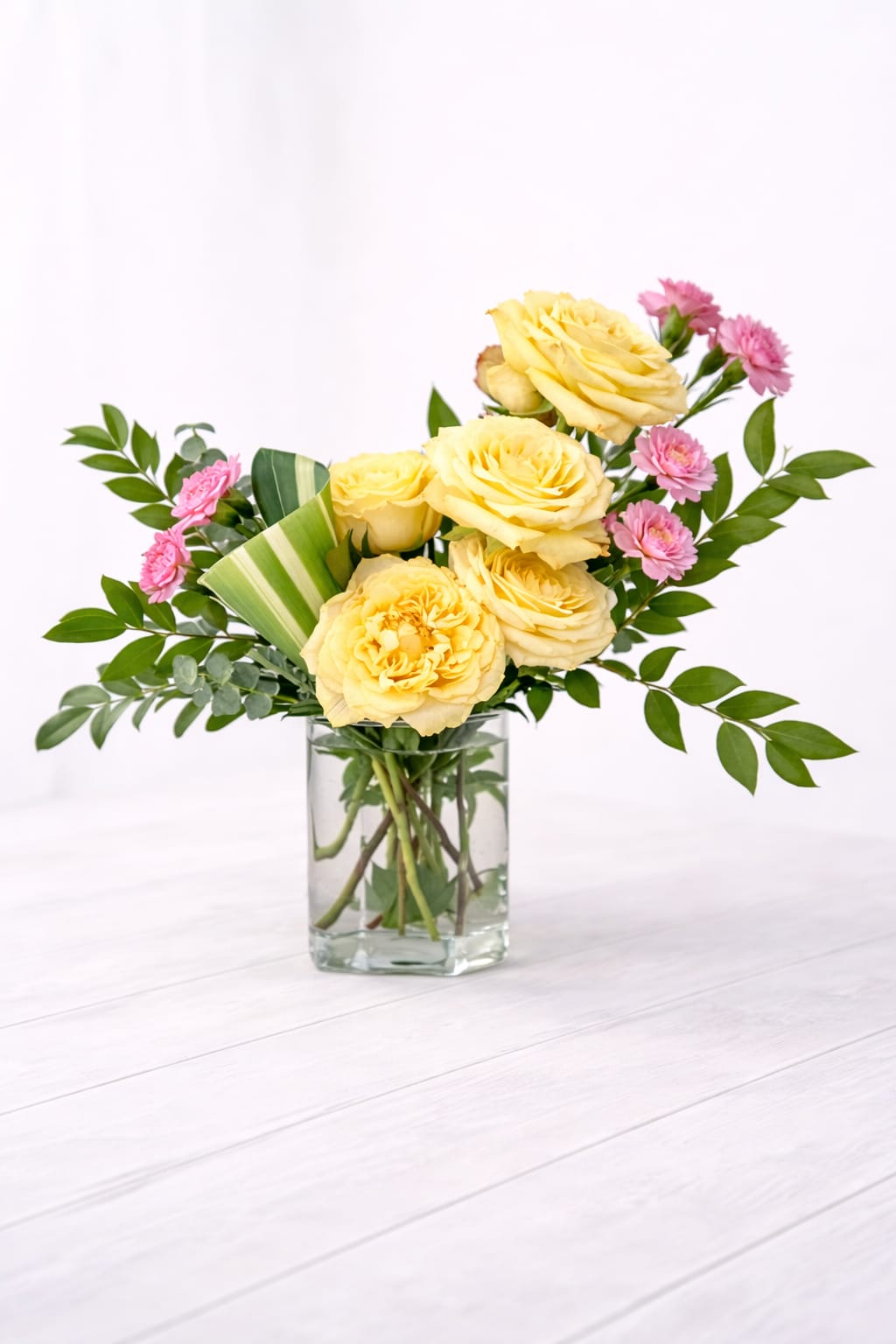 Yellow roses and pink carnations in a clear rectangular glass vase on a light surface
