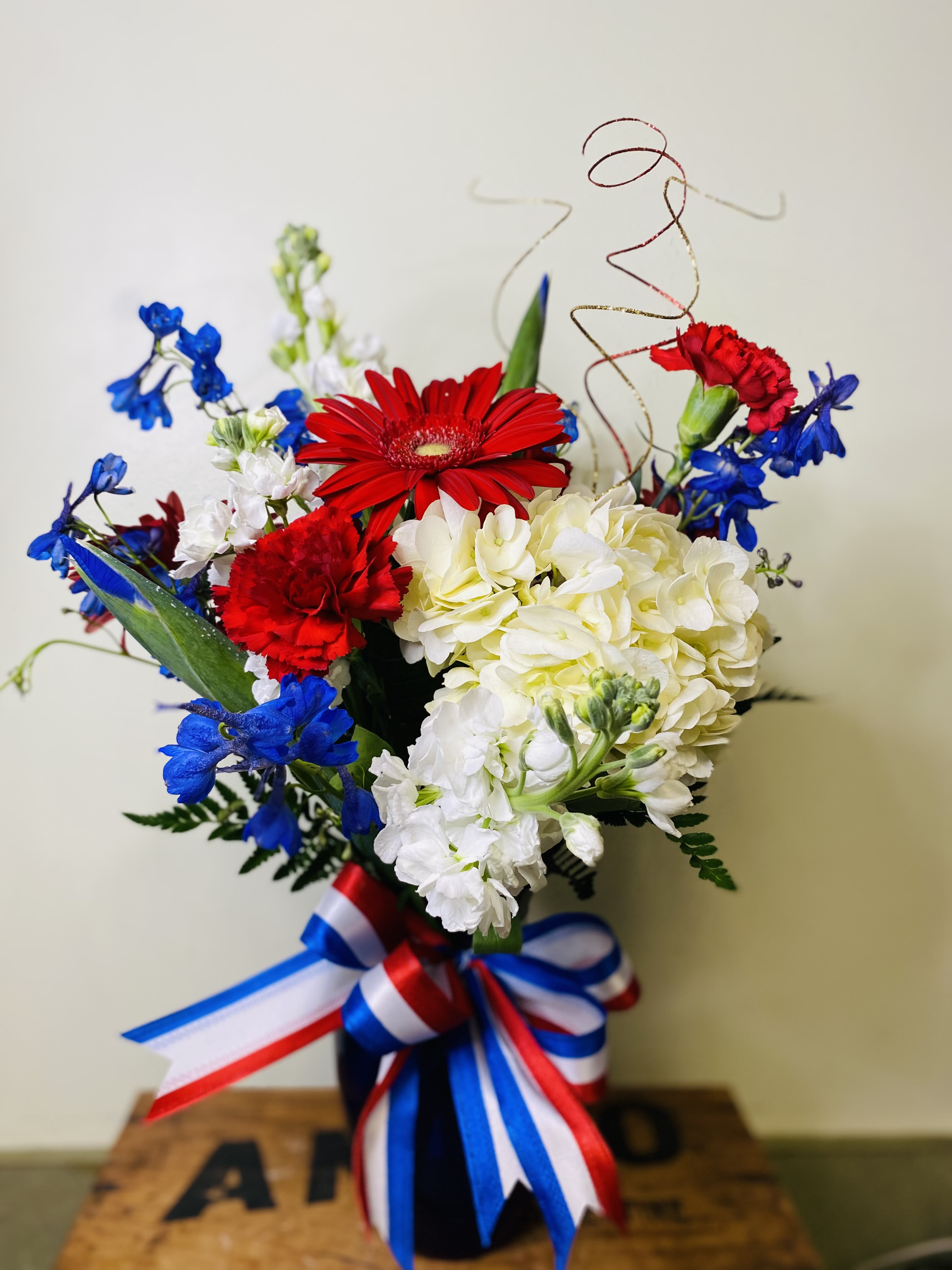 The Firecracker Bouquet - A fresh assortment of our red, white and blue flowers including Iris, Delphinium, Carnation, Stock, Hydrangea and Gerbera. Expertly arranged in a blue vase with a bow this arrangement is perfect for any occasion to show your patriotic colors. 