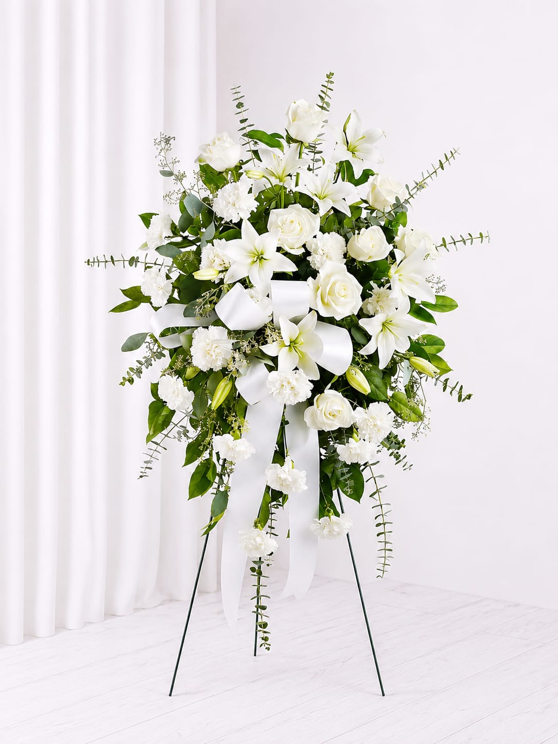 Pure Grace Standing Spray - Elegant in its simplicity, the Pure Grace Standing Sympathy Spray is a timeless tribute designed to offer comfort, peace, and remembrance. This all-white arrangement features graceful lilies, classic white roses, soft carnations, and lush greenery accented with flowing ribbon for a serene and heartfelt presentation. Displayed on a standing easel, this arrangement creates a beautiful expression of sympathy and enduring love during funeral services or celebrations of life.