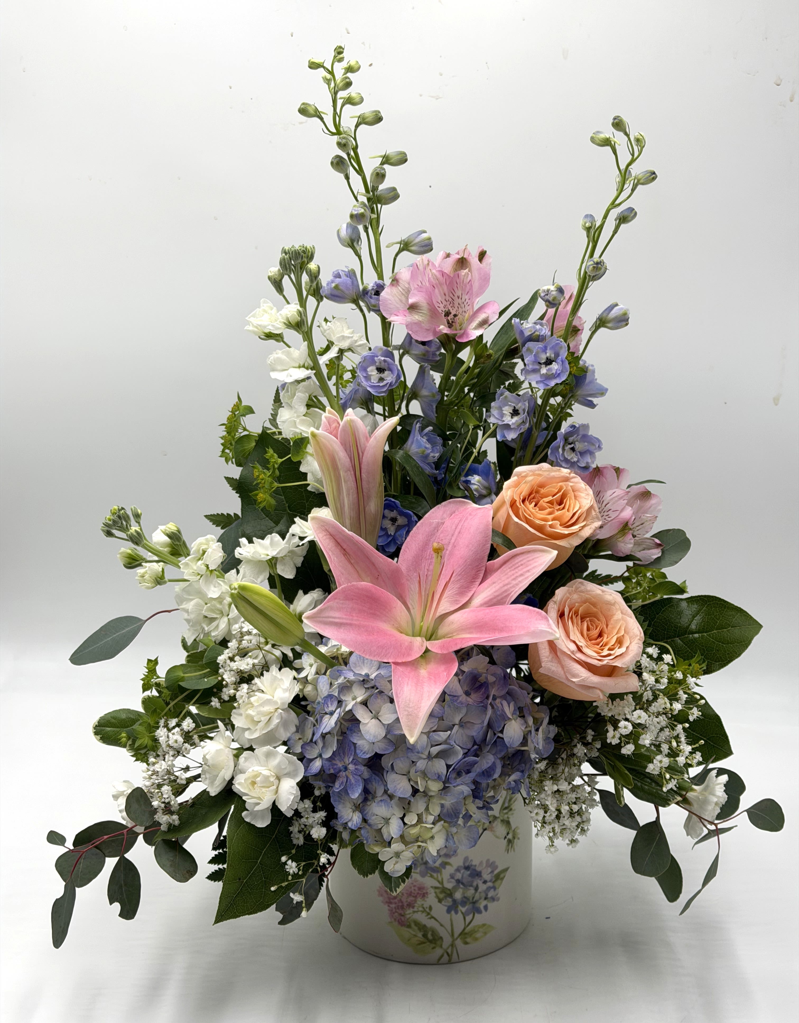 Hummingbirds &amp; Hydrangeas - A soft &amp; elegant garden-style arrangement featuring pink lilies, blush peach roses, blue hydrangea, delphinium &amp; fresh greenery in a charming hummingbird keepsake vase. Perfect for celebrating life's sweet moments with graceful spring beauty!