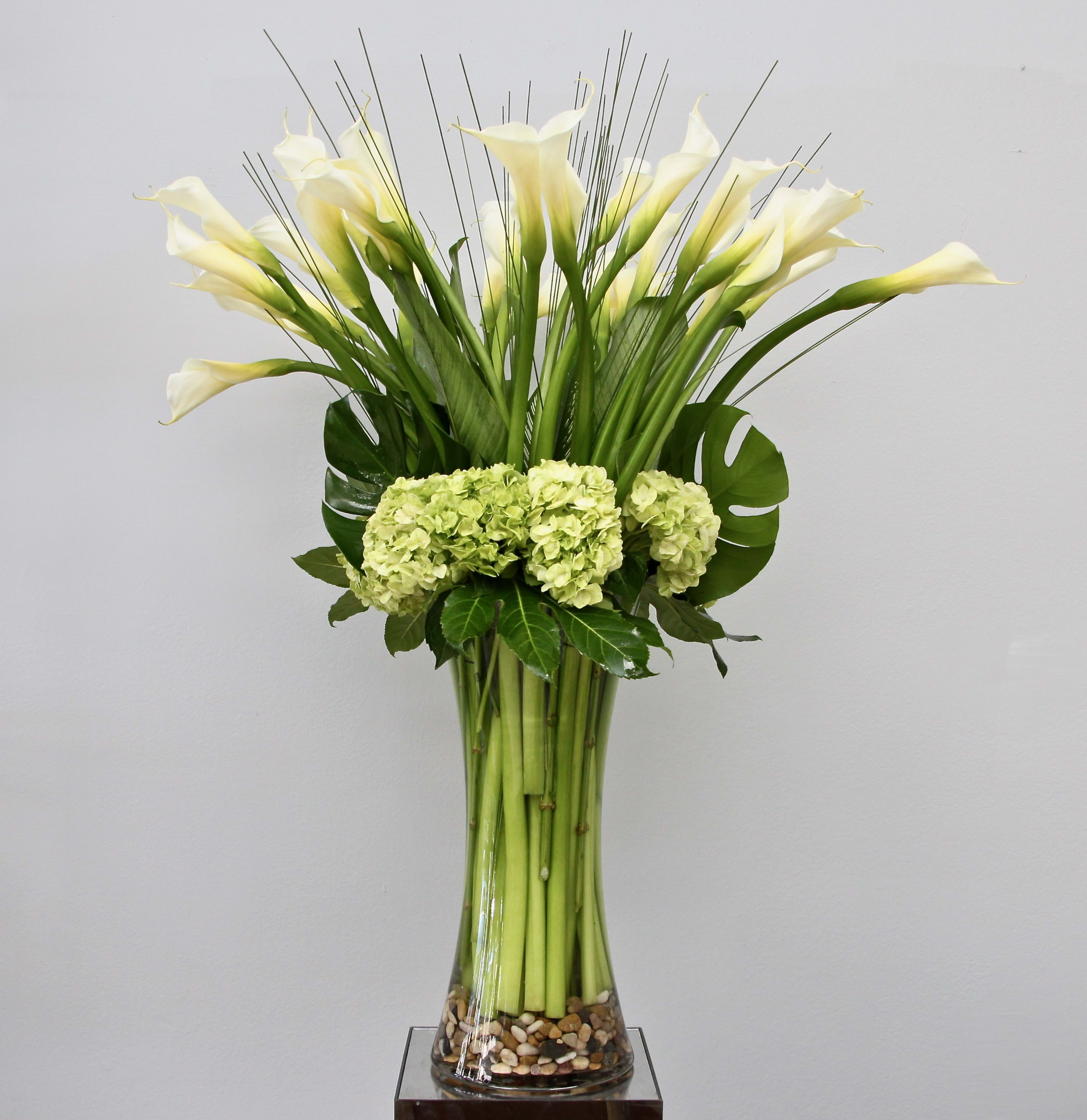 Tall arrangement of white calla lilies and green hydrangeas in a clear glass vase with pebbles