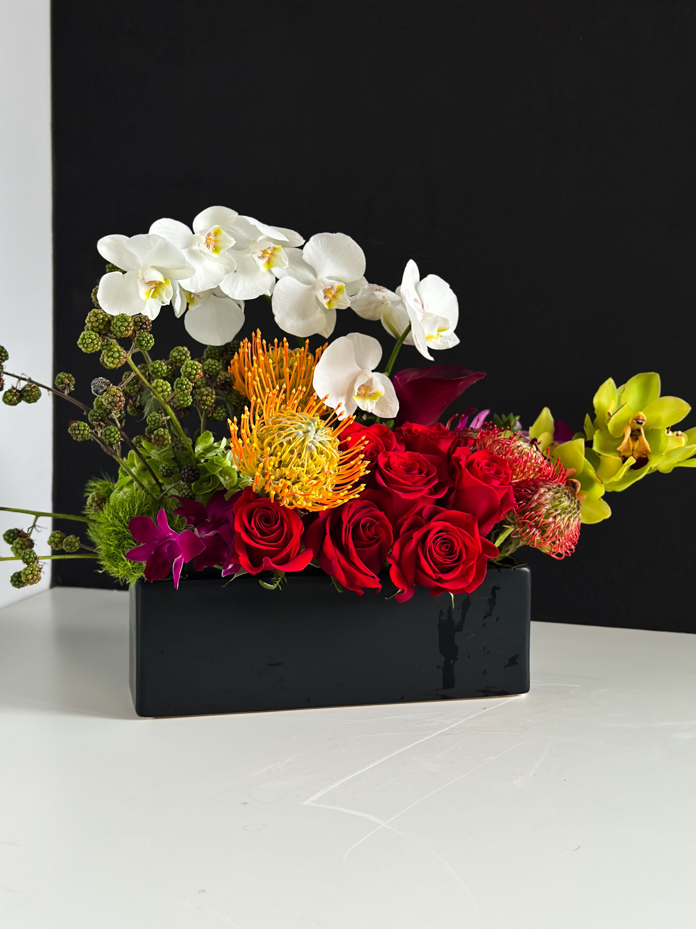 Orchid and Roses - Arching white Phalaenopsis orchids curve gracefully over vivid red roses in a bold, contemporary centerpiece-an anniversary bouquet that also reads beautifully for birthdays, housewarmings, or a "just because" surprise. The dramatic contrast of crisp white, saturated red and pops of orange and purple makes the recipient feel celebrated and truly seen, whether you want to say "I love you," "thank you," or "congratulations."  This low, rectangular black vase holds a lush horizontal composition of Phalaenopsis and Dendrobium orchids, classic roses, orange pincushion-style protea accents, and textured purple and green fillers for depth and movement. The compact, modern silhouette works as a striking mantel or table centerpiece; the arrangement's upscale color palette creates a lasting impression without overpowering the room.  Ideal for someone who appreciates sophisticated, contemporary florals-perfect for anniversaries, romantic gestures, birthdays, or as an elegant housewarming gift. Because of the sturdy vase and structured design, placement is flexible; refresh water as needed and keep flowers out of direct heat to help blooms last.