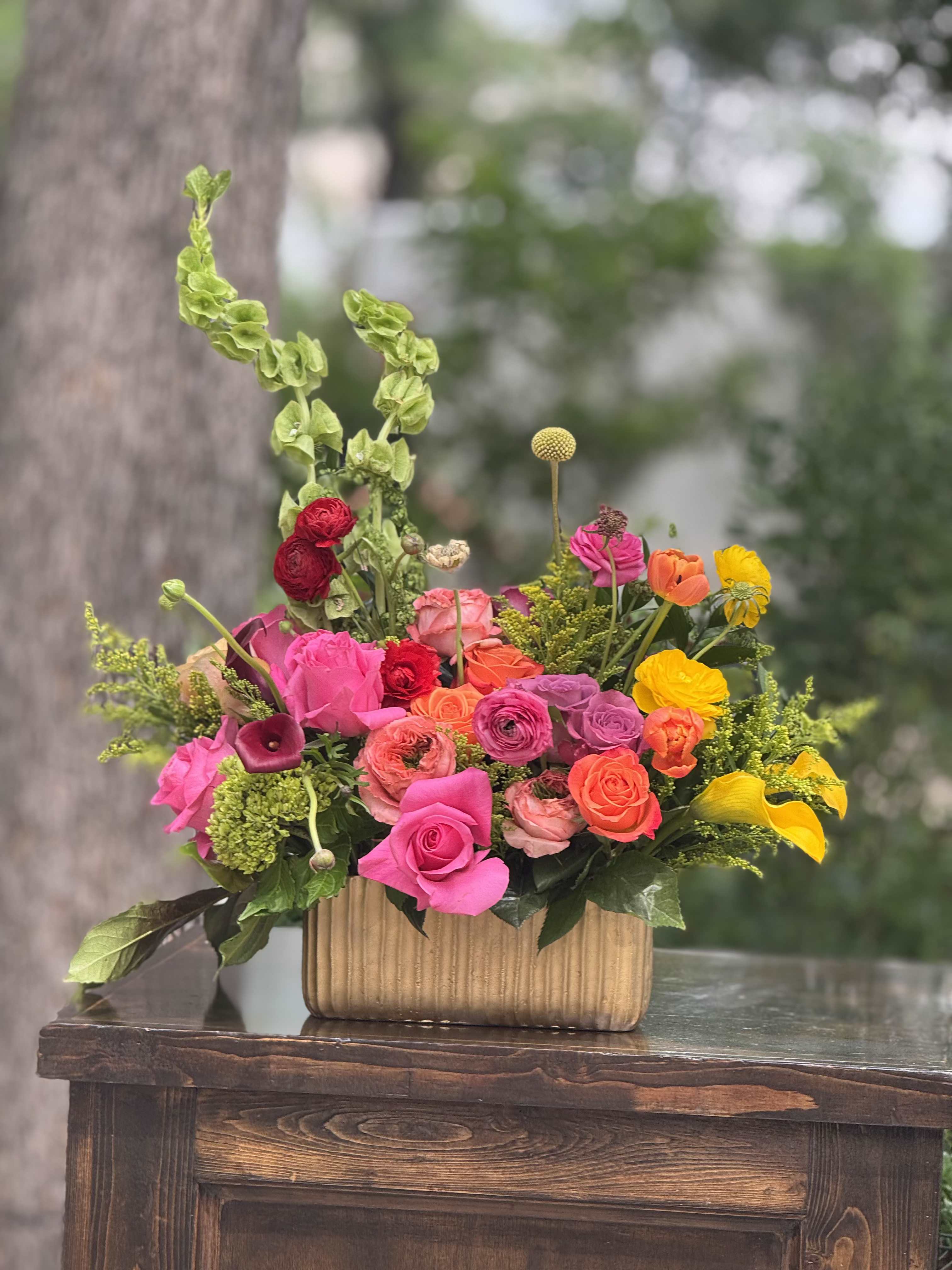 Sunset Garden blooms - A lively, garden-style flower arrangement built around lush roses and ranunculus-perfect as a birthday bouquet, thank-you gift, or bright spring centerpiece. The dense cluster of vivid pinks, coral, orange, yellow and touches of red and lavender creates an uplifting, celebratory feel that will make the person you're thinking of feel warmly noticed and genuinely celebrated.  Rounded rose and ranunculus blooms sit low and close in a ribbed gold rectangular vase, joined by calla lilies, hydrangea accents, taller green textural stems, and airy filler for layered movement and contrast. The low, rectangular design emphasizes color and texture while keeping everything compact and display-ready; the gold container adds a warm, modern shine that complements the palette.  Ideal for sending congratulations, a cheerful "thinking of you," or to brighten a spring gathering-this arrangement arrives arranged and ready to enjoy. Care: keep in fresh water, trim stems periodically, and place away from direct heat to prolong bloom life.