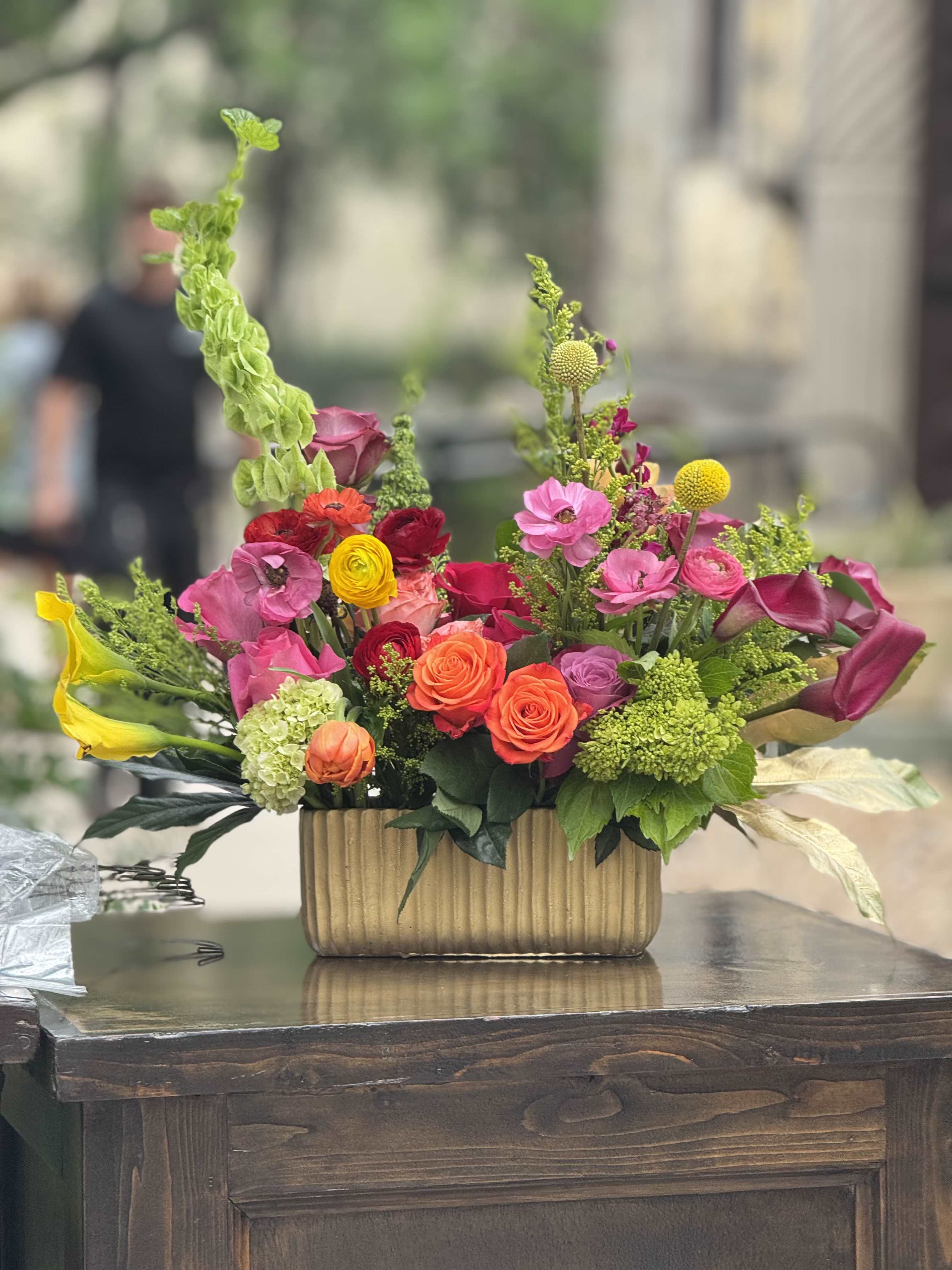 Garden Bloom Luxe - Bright, joyful color pops of pink, coral, orange, yellow and magenta come together in this celebratory mixed bouquet-perfect as a birthday bouquet, congratulations arrangement, or festive everyday gift. The design reads like a modern garden with dramatic sculptural lines: calla lilies extend outward from both sides while tall green accents rise above a dense central cluster, so the person you're surprising will see both movement and abundance at first glance.  A layered mix of roses, ranunculus, hydrangeas and small clustered accent flowers creates varied texture and rounded depth against upright stems. The blooms are arranged in a ribbed gold rectangular vase that adds a polished, upscale finish and a warm metallic contrast to the vivid petals. Sized to make an impression on a table or desk, this arrangement feels lively, cheerful and sure to make someone feel truly celebrated.