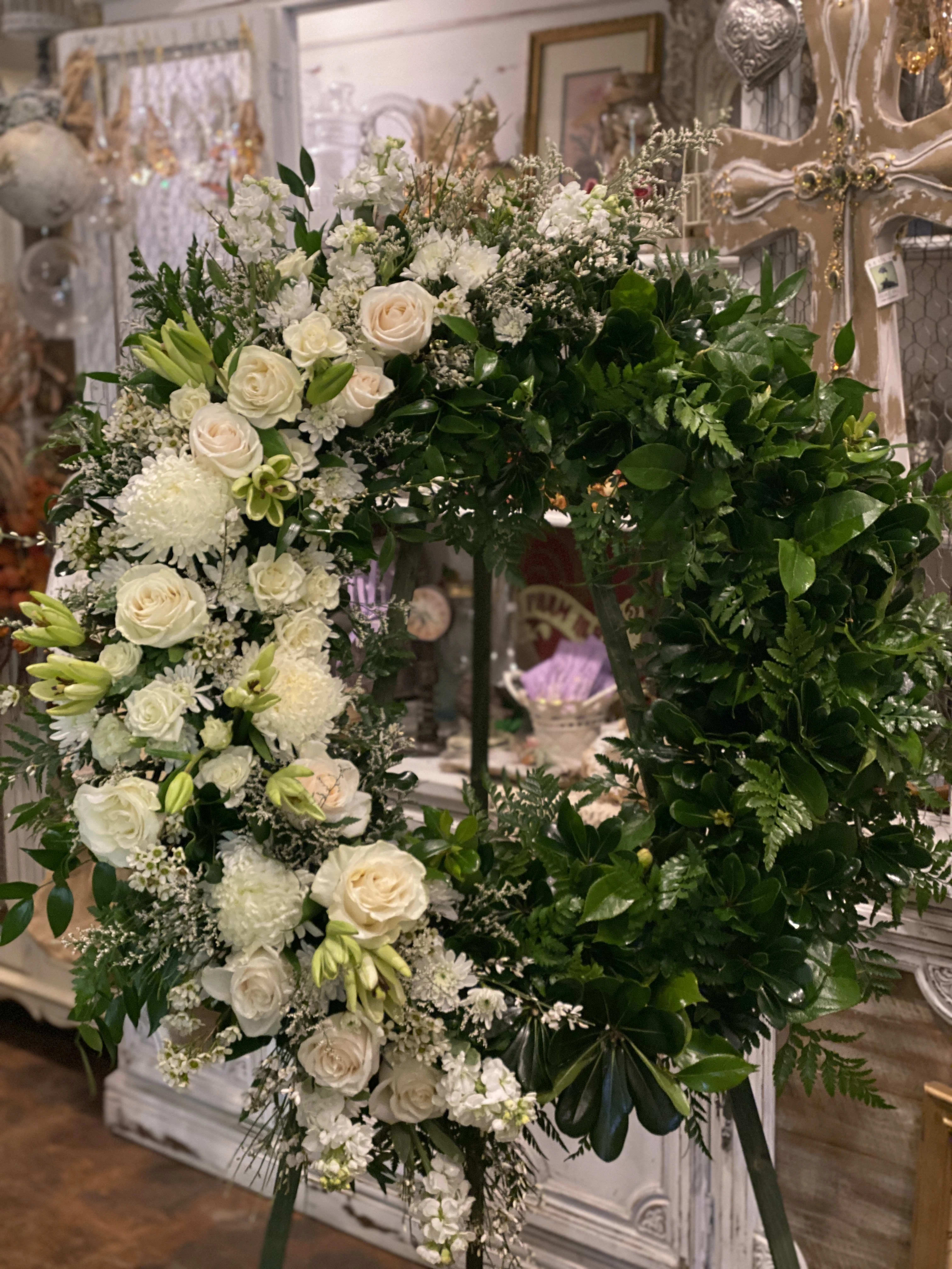  Funeral Spray Wreath - A beautiful wreath adorned with white and ivory roses, lilies, stock and Chrysanthemum mums blooms accented with many greens, and displayed on a green wooden easel. 