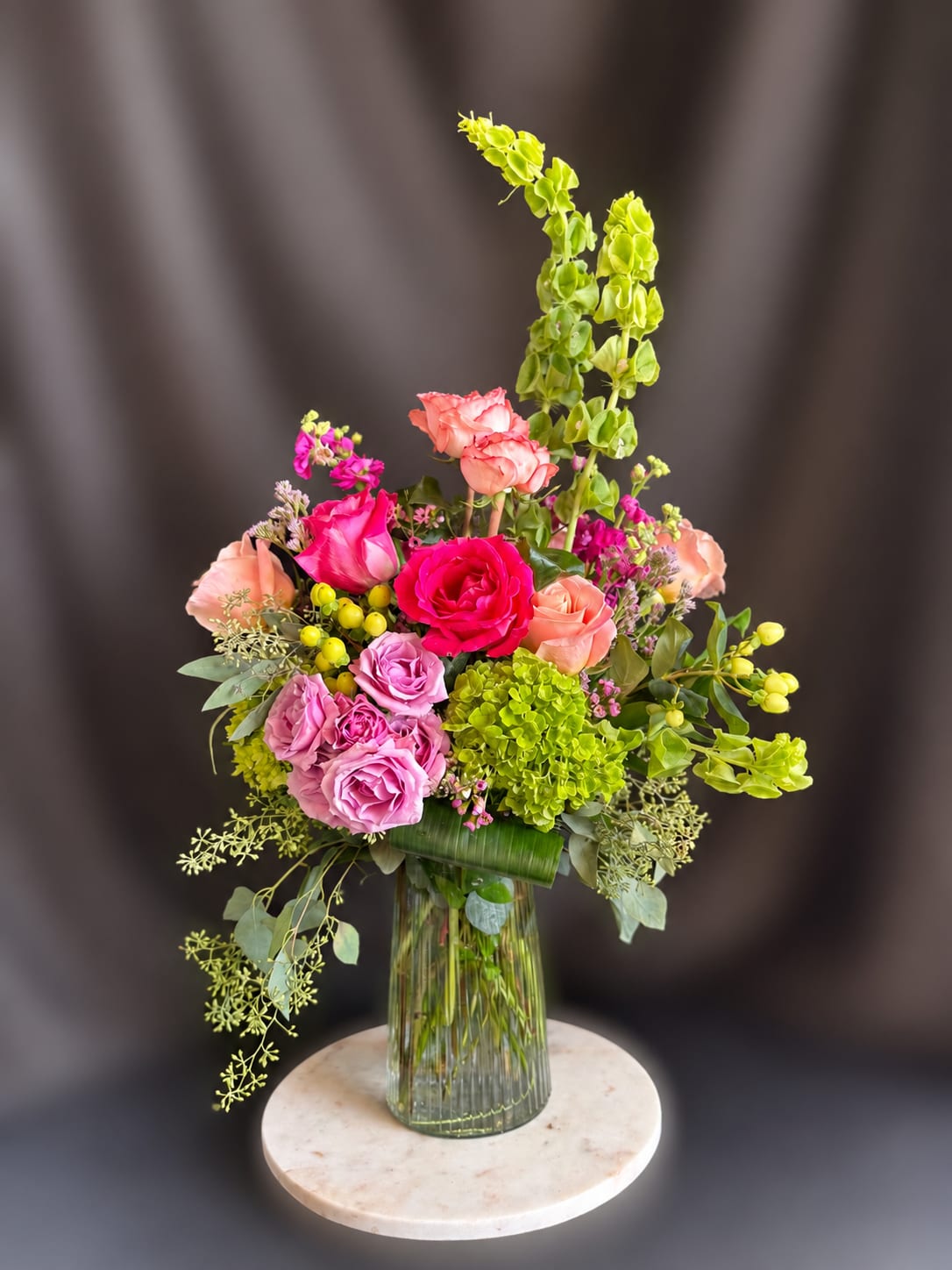 Bells of Eden - Bright pink and blush roses pair with a vivid green hydrangea cluster in a lively garden-style bouquet perfect for a birthday bouquet, thank-you gift, or spring celebration. Tall lime-green snapdragon spikes and an upright green flowering stem create elegant vertical movement, while peach, lavender and magenta accents - plus small berry-like filler - add texture and joyful contrast so the recipient feels genuinely celebrated.  Arranged in a clear glass vase with a decorative leaf wrap, this floral arrangement mixes romantic softness and fresh, clean lines for an airy yet abundant look. The palette of pinks, greens, white and purple reads cheerful and polished; the design rises upward but spreads into a full, rounded base for visual balance. Ideal for everyday gifting when you want someone to feel seen, it arrives ready to display - simply trim stems occasionally and refresh the water to keep blooms looking their best.