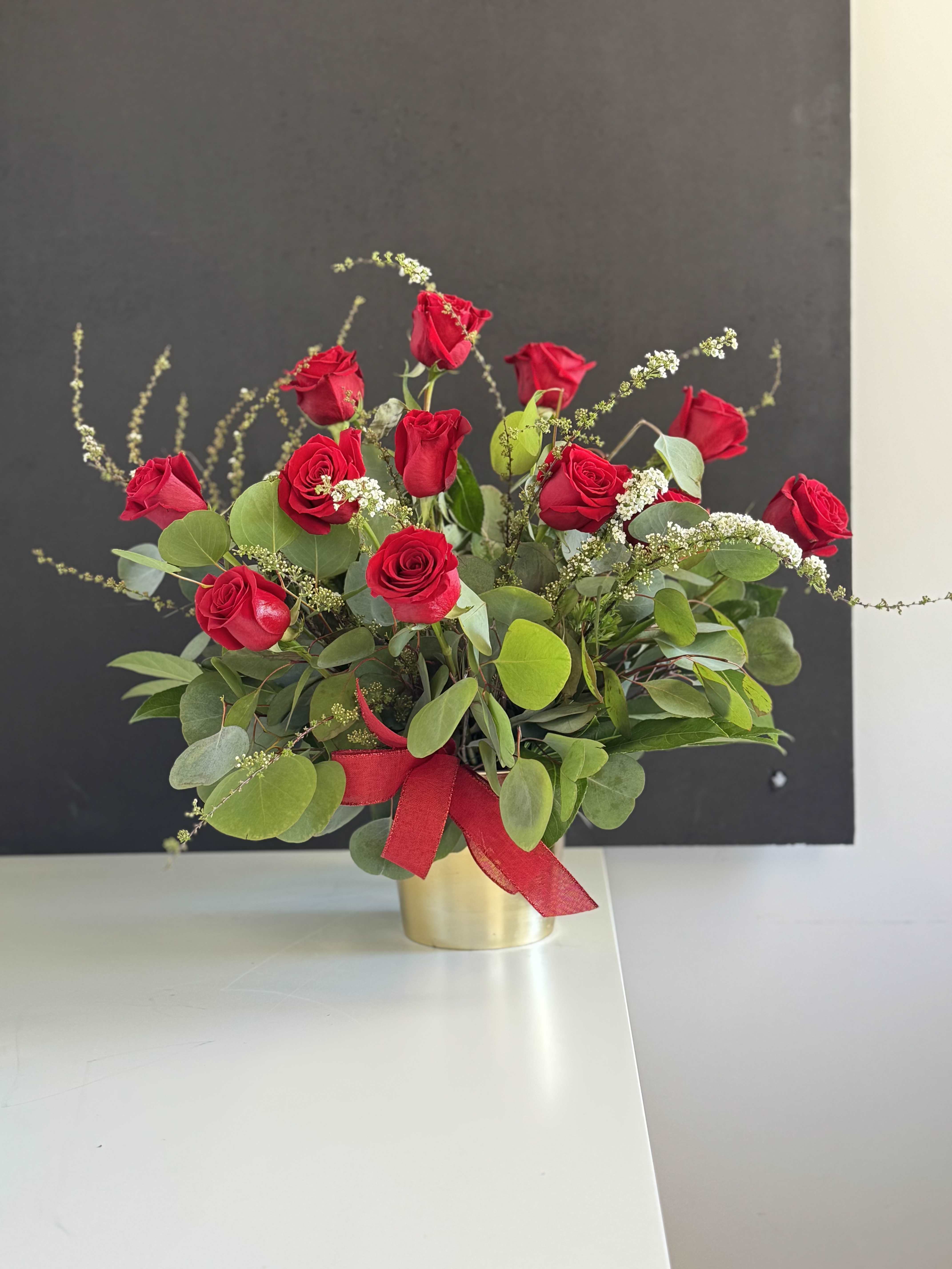 Tribute 12 Roses - Velvety red roses clustered with airy white filler and soft, round eucalyptus create a romantic flower arrangement perfect for an anniversary, Valentine's Day, or to say "I love you" any day. The red ribbon tied around the base and the warm gold vase give the piece a polished, celebratory feel-she'll notice the rich color and graceful movement of the blooms the moment it arrives.  This arrangement is built in a low, rounded composition with blooms set at slightly varied heights for a naturally loose yet refined silhouette. Multiple red roses are the focal point, balanced by delicate white filler for lightness and abundant, rounded eucalyptus-like foliage for texture and green contrast. The flowers come presented in a small gold container adorned with a red ribbon, creating an elegant keepsake vessel that complements the classic color palette.  Ideal for the person you want to make feel cherished-whether marking a milestone, saying thank you, or surprising a loved one-this piece arrives ready to display. Standard care: keep in fresh water, trim stems and refresh water every 2-3 days to extend bloom life.