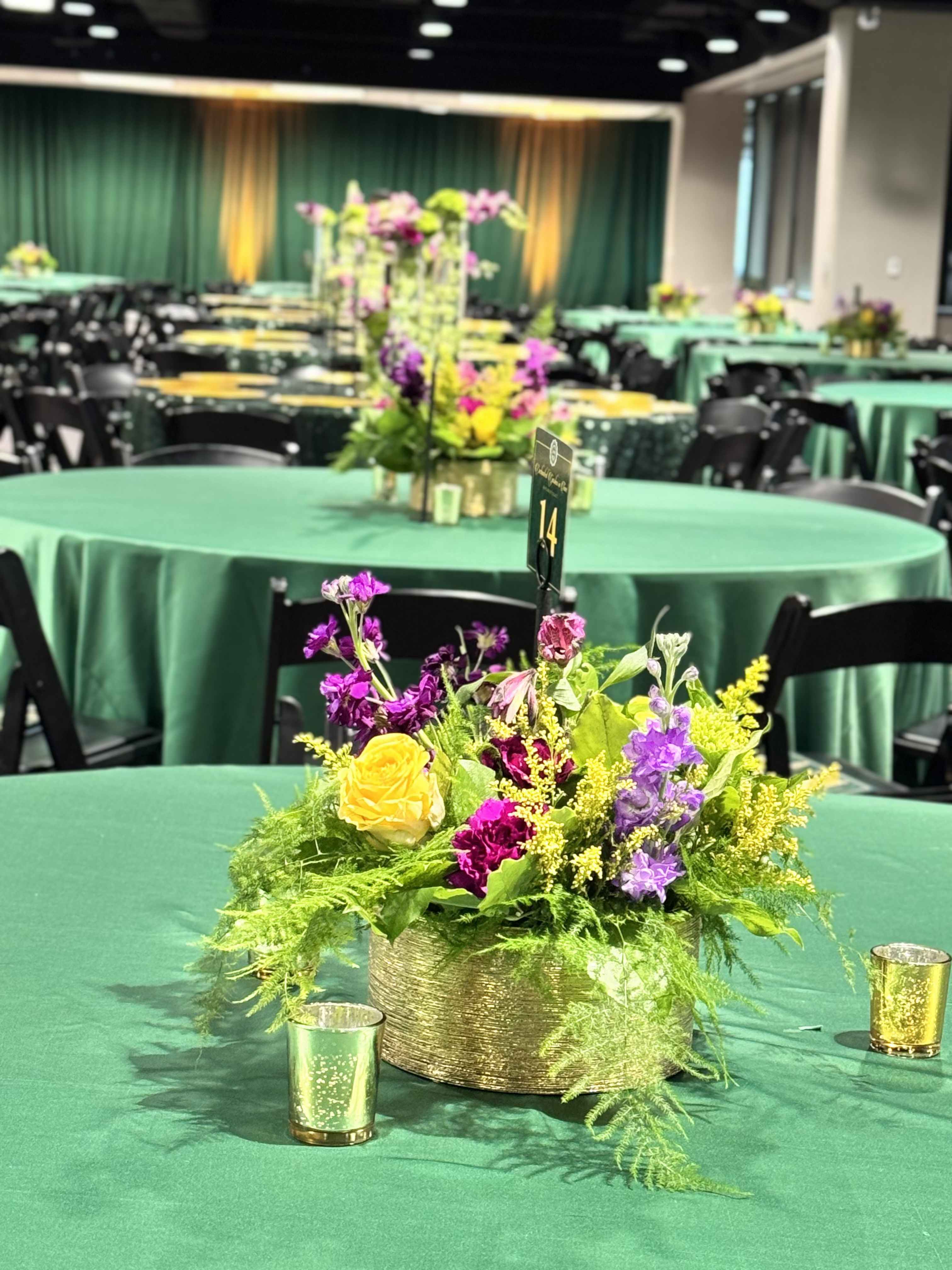 Green Lux - A low, full centerpiece in a textured gold round container brings radiant spring energy to a wedding reception or celebratory banquet. This table centerpiece for a reception or large event mixes yellow, magenta, purple and lavender blooms with airy green filler so the person you're honoring will feel uplifted and celebrated the moment they sit down. The vibrant color mix and polished gold vessel read as festive yet refined-perfect for spring weddings, anniversary dinners, or any joyful gathering. The arrangement combines roses, carnations, stock and complementary seasonal flowers clustered tightly for a lush, garden-fresh silhouette. Set low to sit comfortably on round tables, the centerpiece measures to coordinate with standard table settings and allows clear sightlines across the table; pictured on a green tablecloth, it pairs beautifully with gold accents like a round container, table number sign and glass votive candle holders. The textured gold container adds warmth and a luxe touch, while the mix of bright and soft hues creates depth and movement across the display. Designed for event tables, this centerpiece works as a coordinated set across multiple banquet tables to create a cohesive reception look that feels both festive and polished. 
