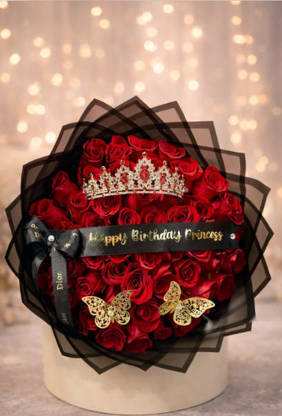Royal Birthday Princess Bouquet - A dense dome of velvety red roses crowns this birthday bouquet, arranged in a chic round hat box for a bold, romantic presentation - perfect as a birthday bouquet for a girlfriend, wife, or anyone who deserves to feel like royalty on their day. The arrangement reads glamorous and indulgent at a glance: layered black sheer wrapping and a black satin ribbon printed "Happy Birthday Princess" announce the occasion while a jeweled crown perched atop the roses creates a celebratory focal point that says "you are cherished."  Rich red roses are tightly packed to form a lush, sculpted silhouette, accented near the base by two metallic gold butterfly ornaments that add movement and sparkle. The black hat box keeps the design compact and gift-ready; finishing touches include an additional bow with printed text and a birthday message ribbon for personalization. This bouquet is ideal for romantic anniversaries and milestone birthdays where the goal is to make her feel truly seen - delivery-ready in its hat box format and designed to arrive as a show-stopping, long-lasting expression of admiration.