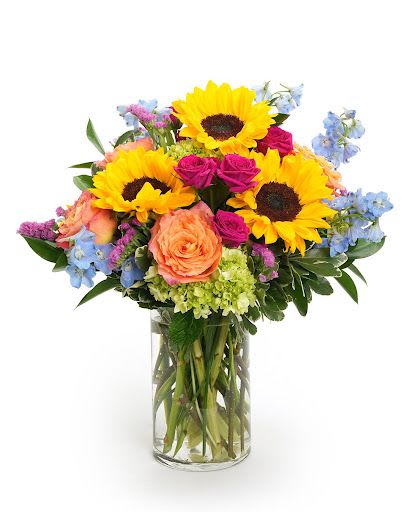 Arrangement of yellow sunflowers, pink roses, blue delphinium, and green hydrangeas in a clear glass vase.
