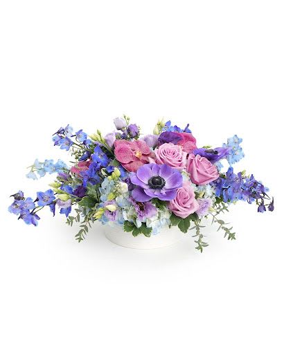 Low arrangement of purple anemones, lavender roses, and blue flowers in a white bowl.