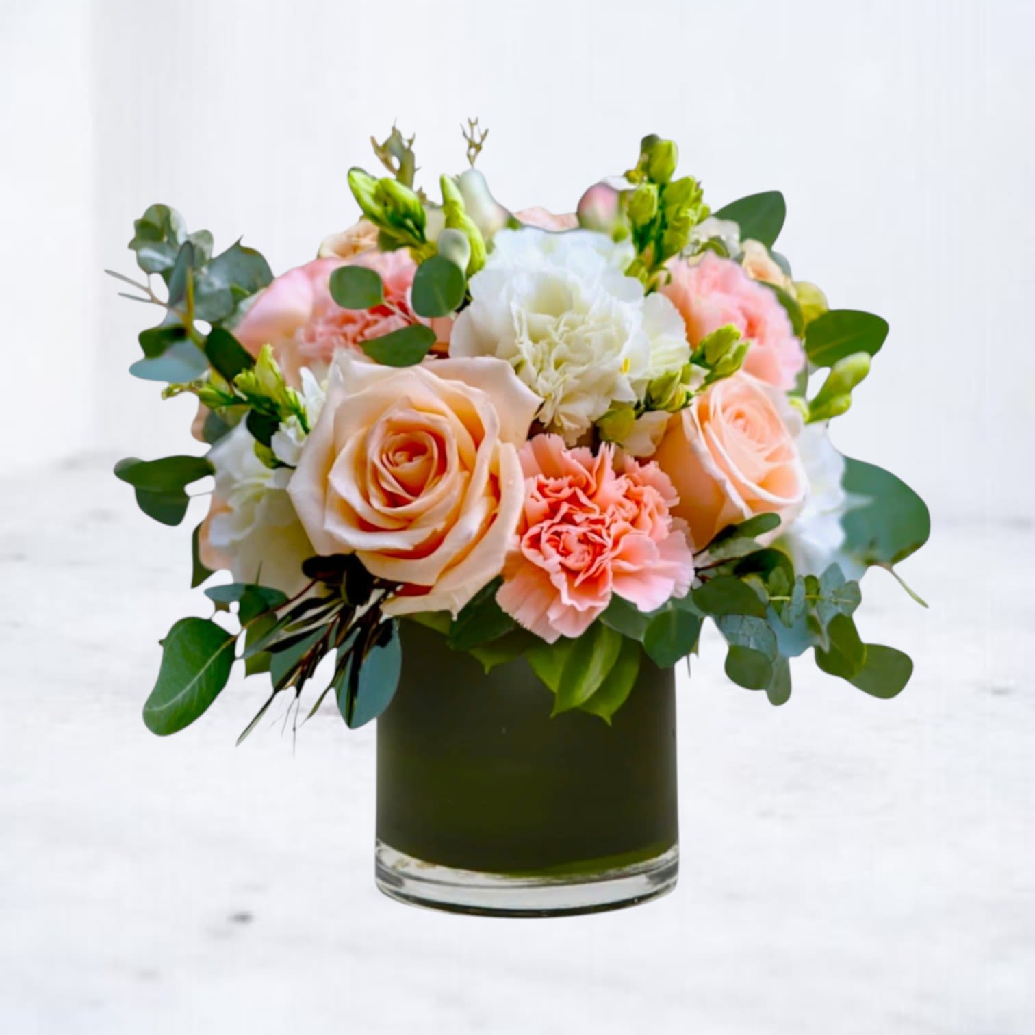 Short arrangement of peach roses and pink and white carnations in a green glass cylinder vase