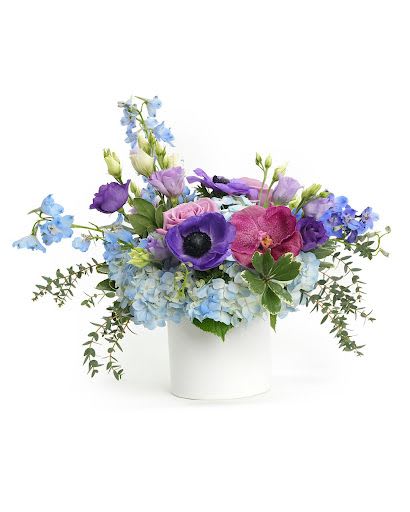 Low arrangement of blue hydrangea and purple blooms in a white cylinder vase