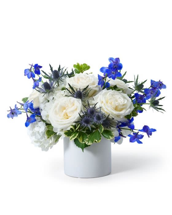 Low white and blue flower arrangement with roses in a white cylinder vase