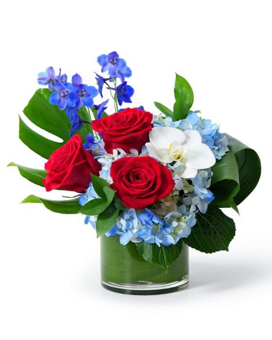 Short arrangement of red roses, blue flowers, blue hydrangea and a white orchid in a glass vase