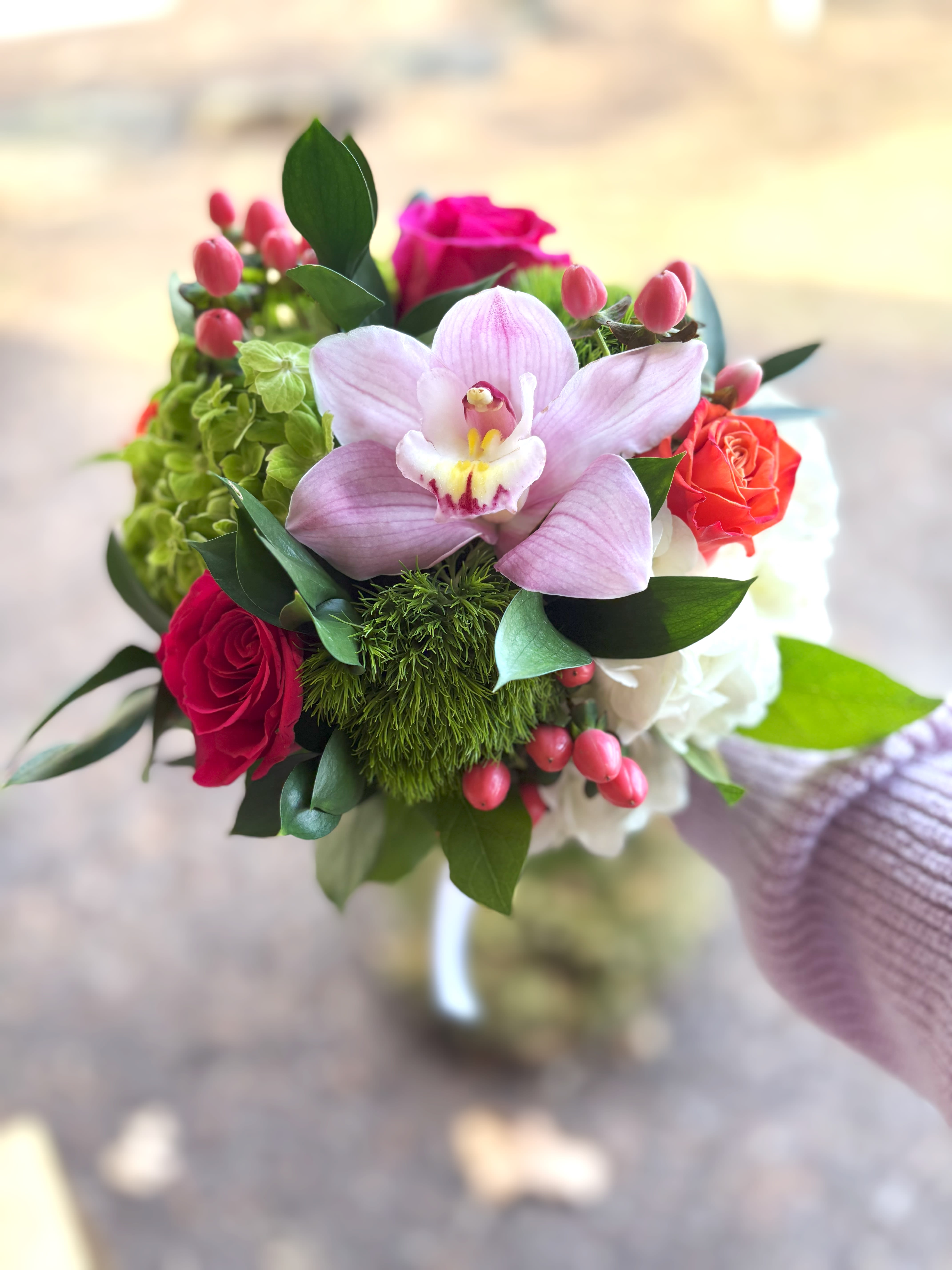 Handheld bouquet with pink orchid, red and orange roses, green blooms, and pink berries