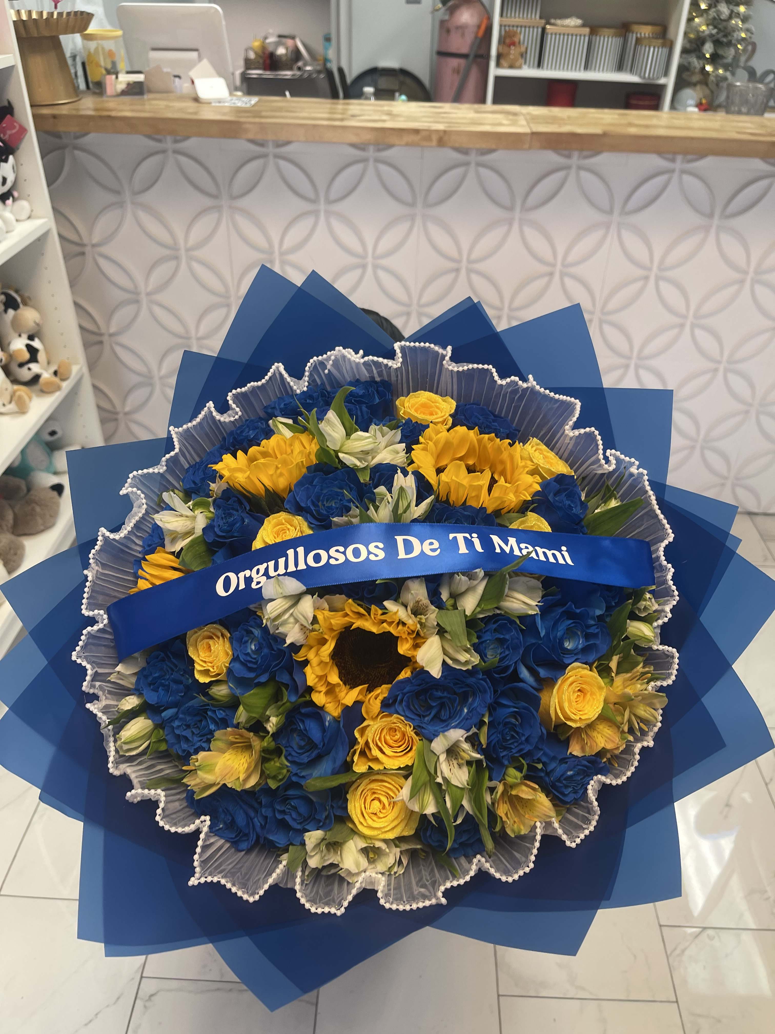 Blue and yellow bouquet with a ribbon reading "Orgullosos De Ti Mami"