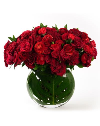 Low round arrangement of red roses and ranunculus in a clear glass vase lined with a large green leaf.