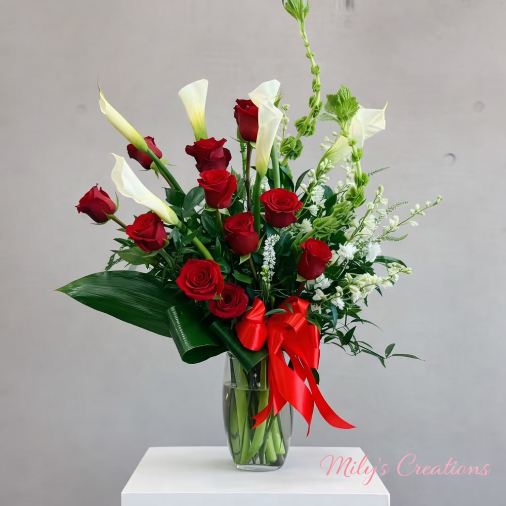 Tall arrangement of red roses, white calla lilies, and green Bells of Ireland in a glass vase with a red bow