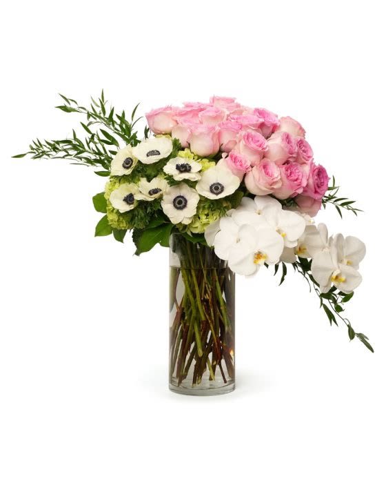 Asymmetrical arrangement of pink roses, white anemones, and orchids in a tall clear glass cylinder vase.