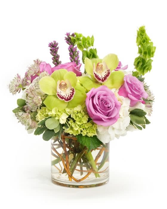 Short arrangement of green orchids, pink roses, and white blooms in a clear glass cylinder vase