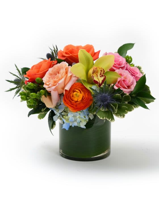 Low arrangement of orange and pink roses, green orchid, and mixed blooms in a clear glass cylinder vase.