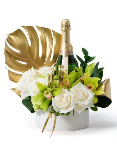 White roses and green orchids arranged around a champagne bottle with gold accents in a white container