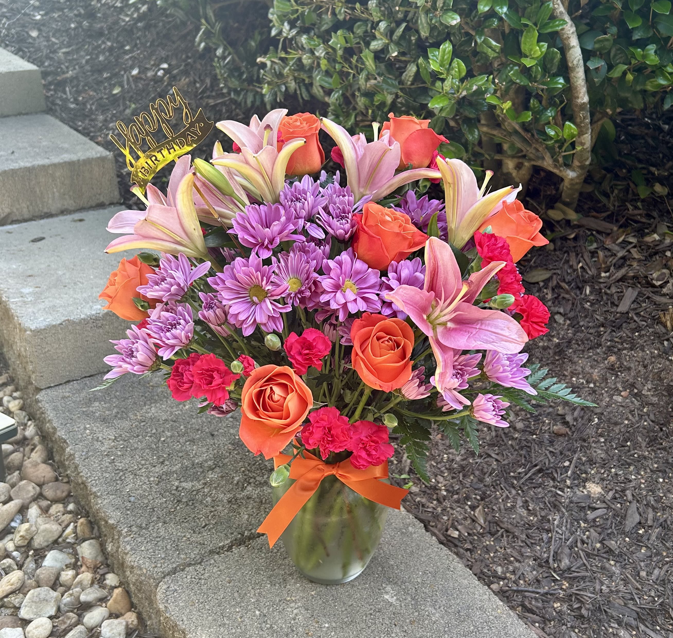 Mixed bouquet of pink lilies, orange roses, and purple daisies in a glass vase