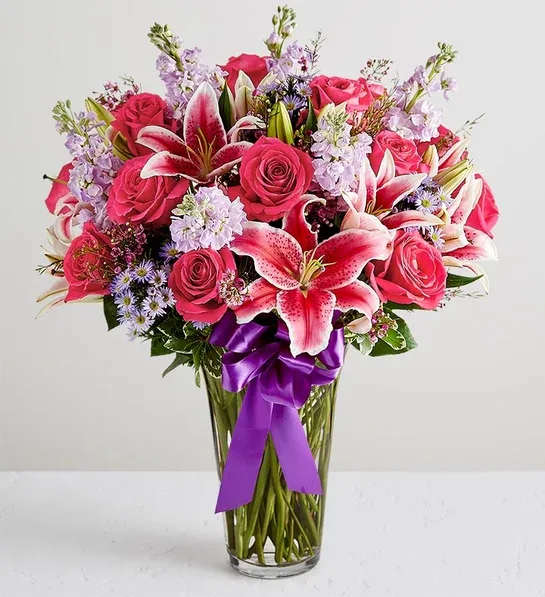 Pink roses and lilies in a clear glass vase with a purple ribbon