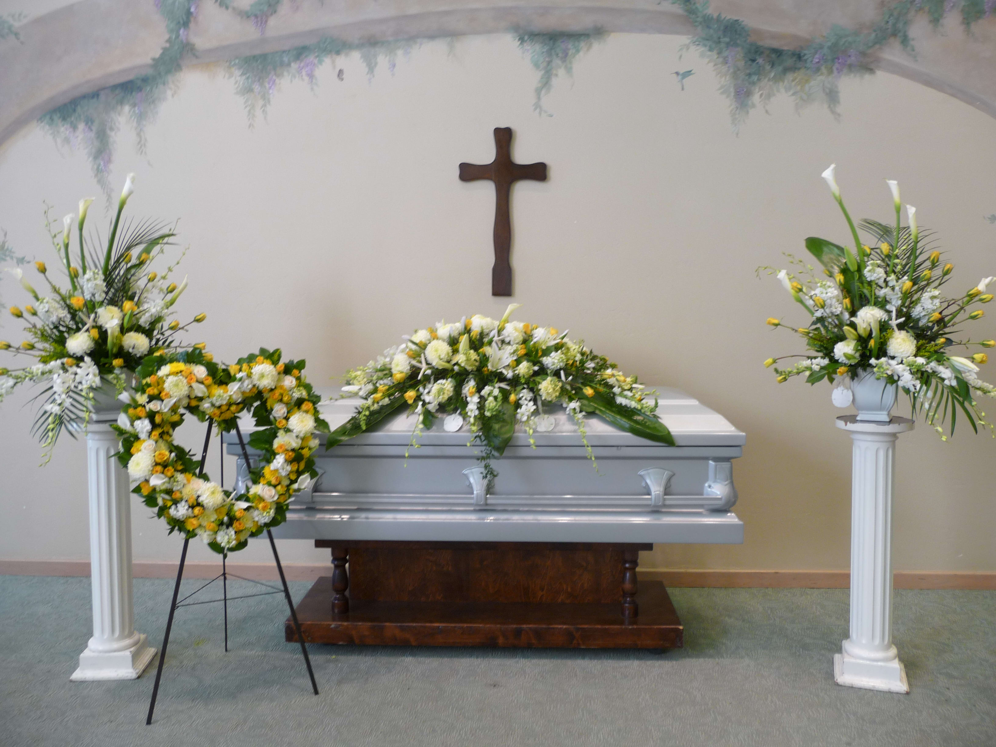 Funeral casket with white and yellow floral sprays, heart wreath, and pedestal arrangements.