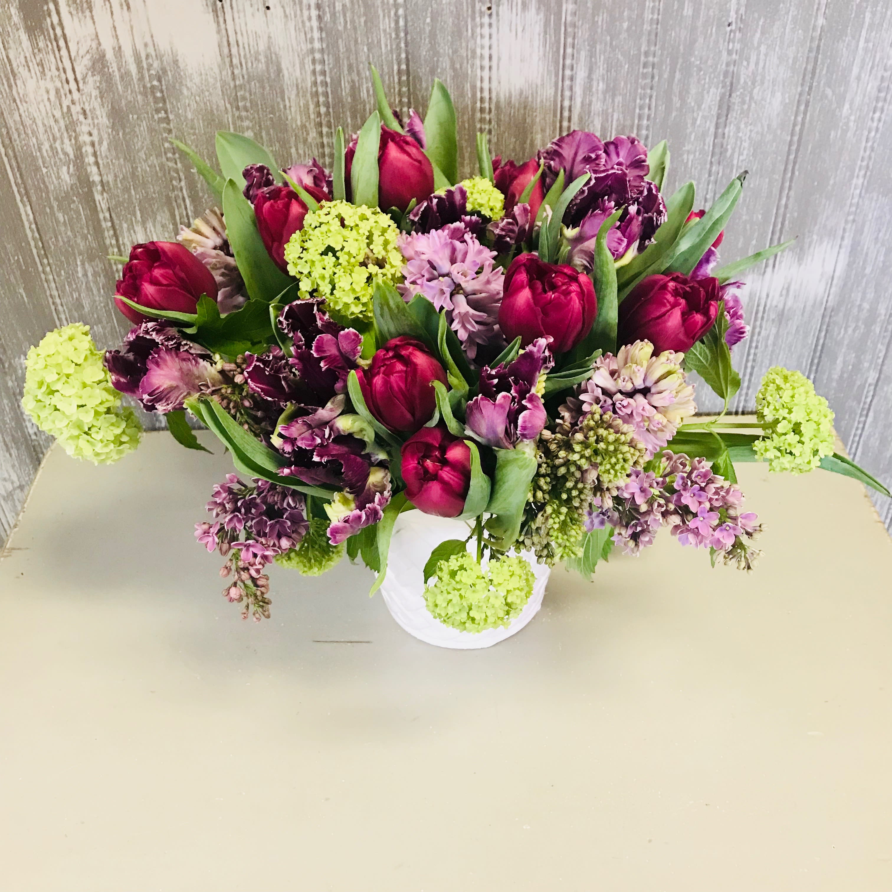 Pixie Spring - A beautiful arrangement with novelty tulips, hyacinths, lilac and viburnum in a white lattice vase in shades of plums and lavenders.