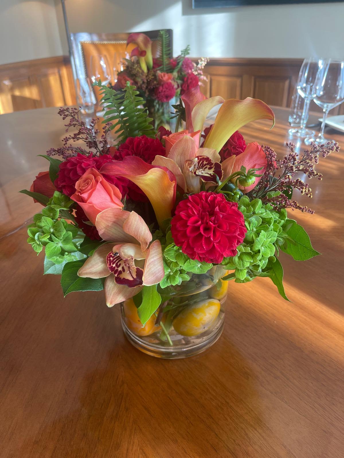 Low glass vase of red dahlias, peach roses, calla lilies, orchids, and green blooms on a wood table
