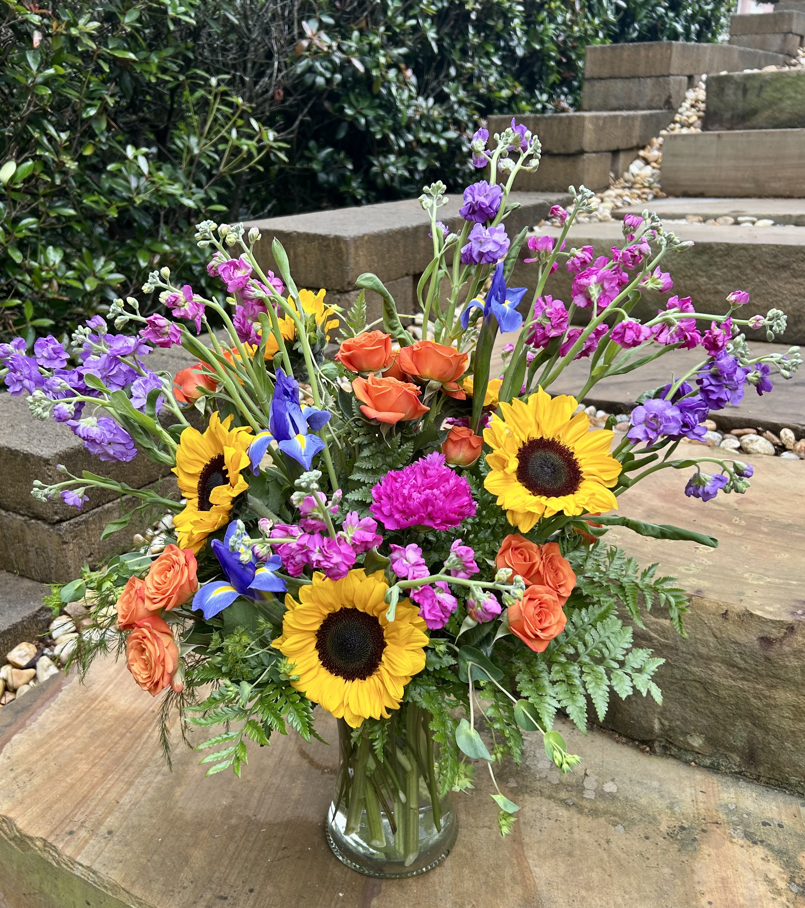 Colorful mixed bouquet in a clear glass vase with sunflowers, roses, and purple blooms