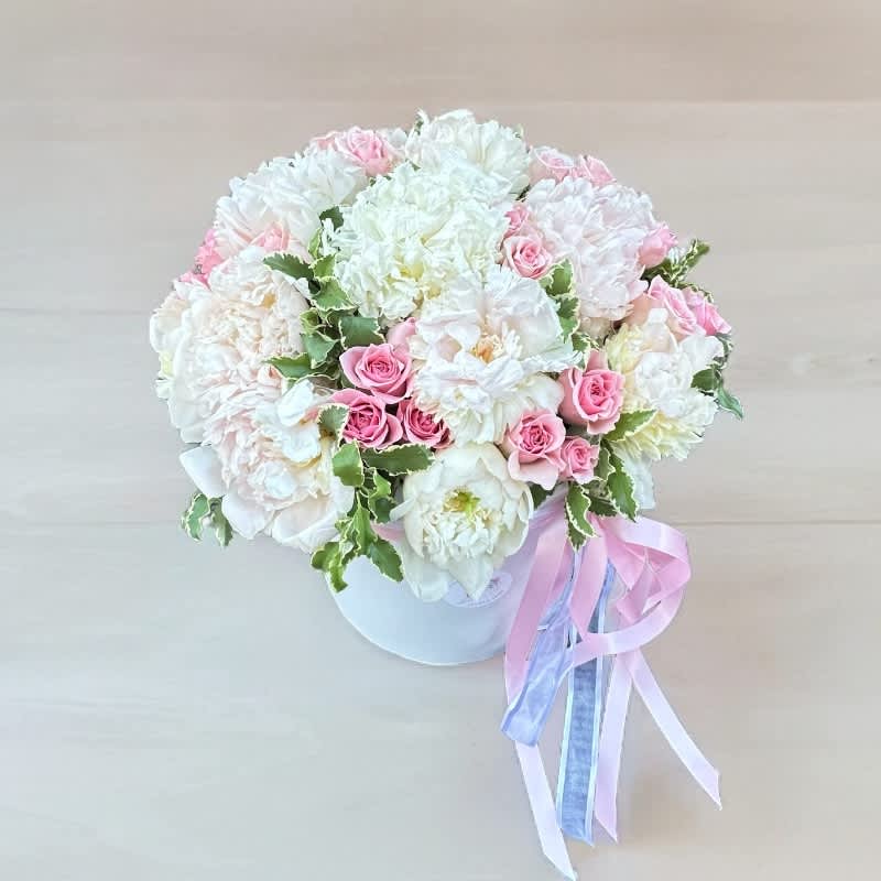 Round bouquet of white and pink flowers in a white box with ribbon tails