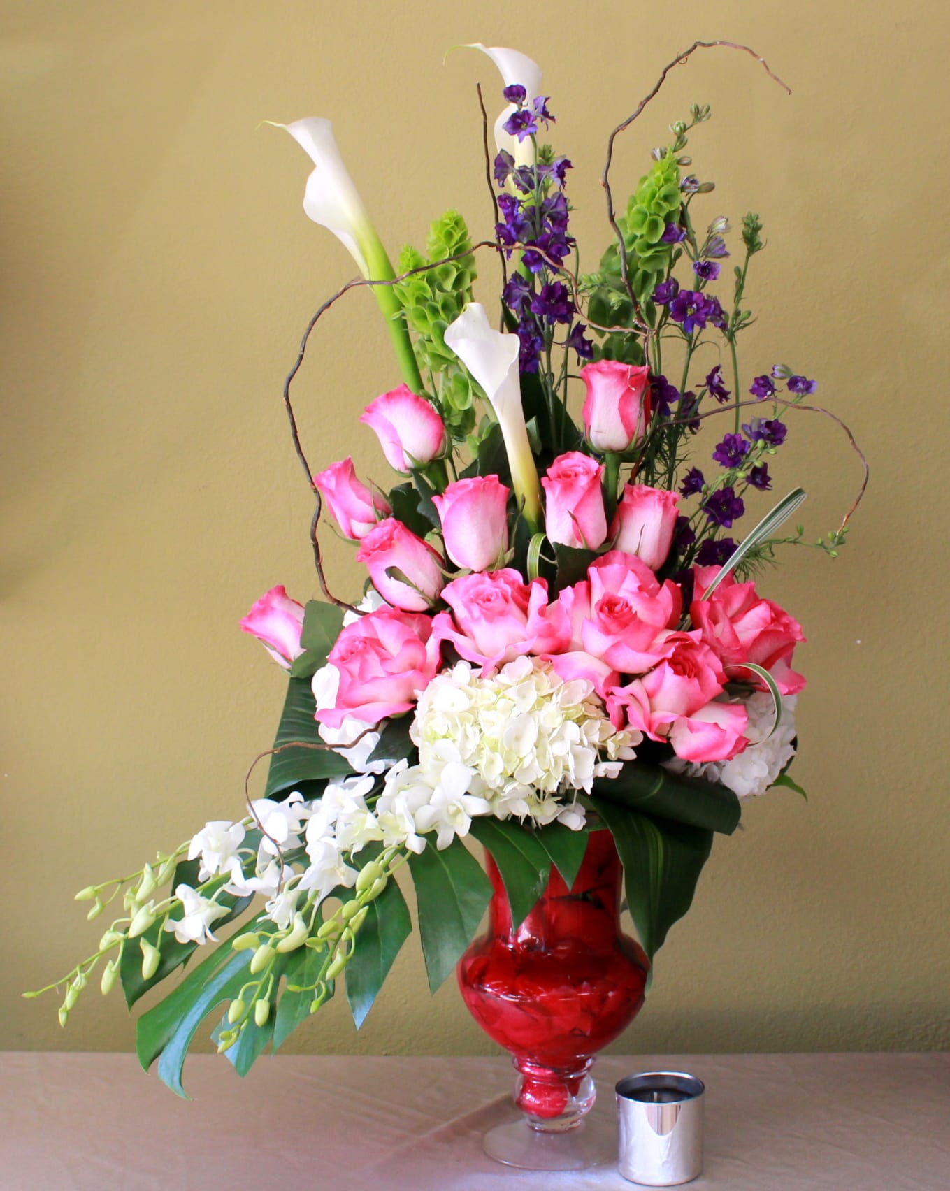 Love Me Lots - white calla lily's, curly willow,  green bells of Ireland , purple larkspur pink long stem roses ,white  hydrangeas and white  dendro orchids in a elegant tall clear vase with red petals inside. 