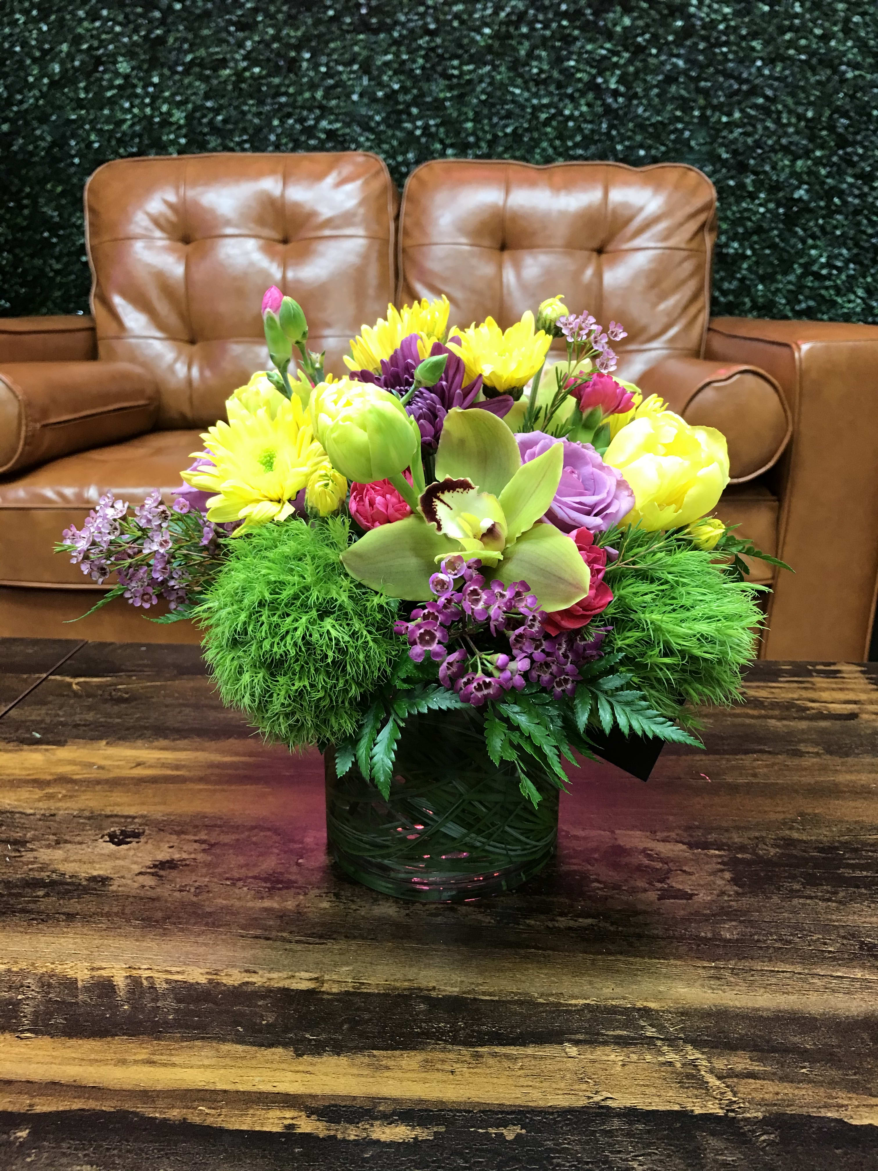 Low glass vase of yellow mums, purple blooms, green dianthus, and an orchid on a wooden table