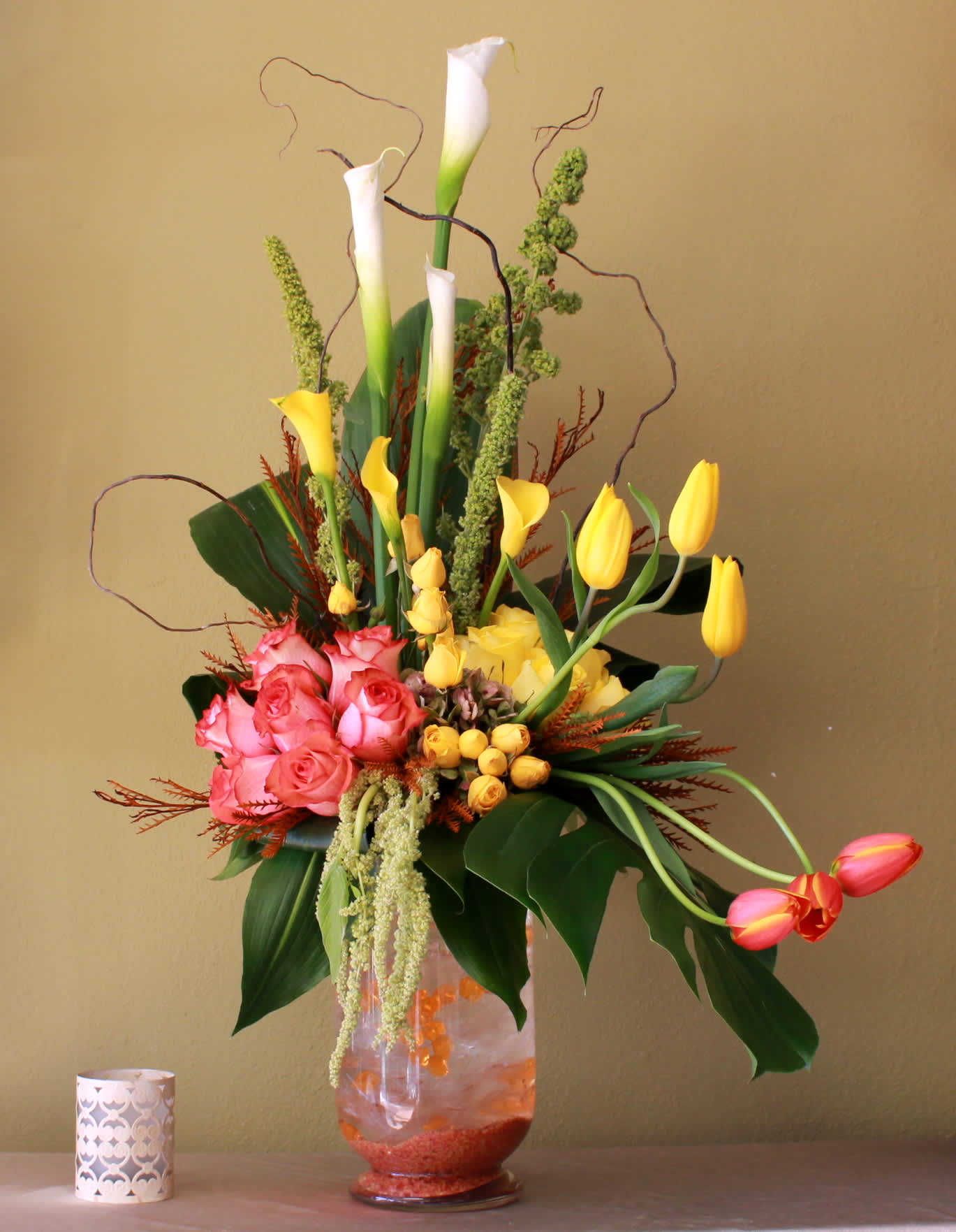 Island Escape - White and yellow calla lily's, yellow and orange tulips, yellow and orange long stem roses, aspidistras leaves and curly willow and other elegant greens in a tall and clear cylinder vase.