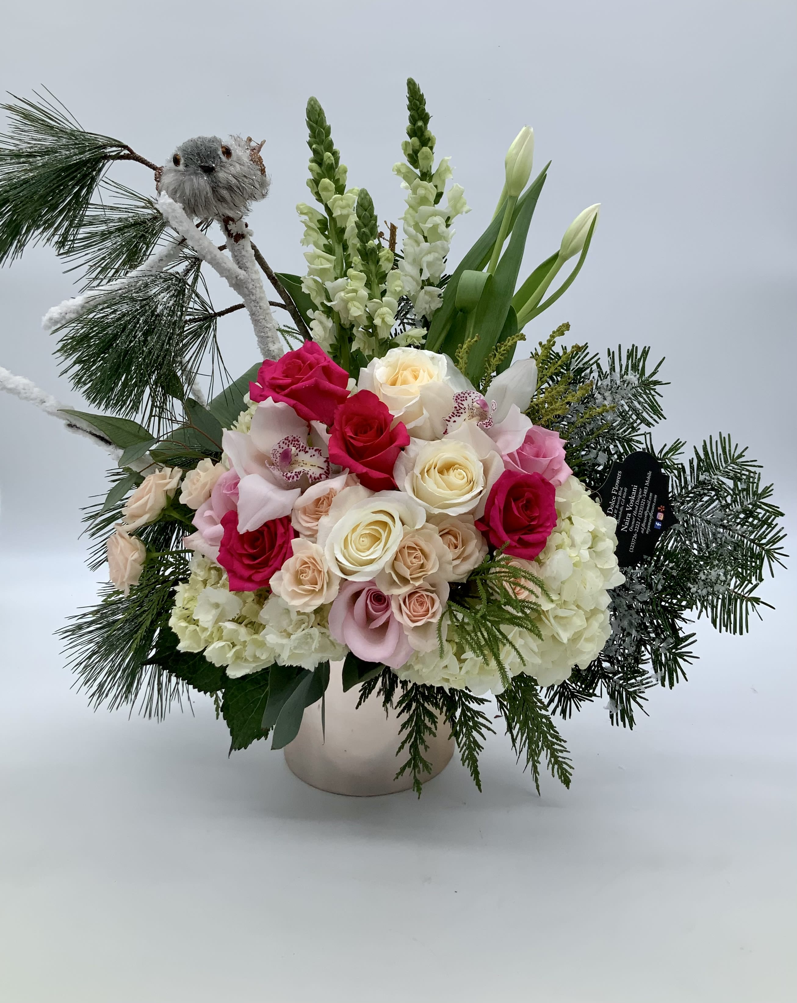 Winter Dreaming  - Perfect for any winter occasion. Made with pink toned flowers, snowy flocked branches, and fragrant Christmas greens. All arranged in a ceramic and metallic rose gold cylinder vase. The arrangement also features a snow covered little bird perched on top of the branches overlooking the flowers. 