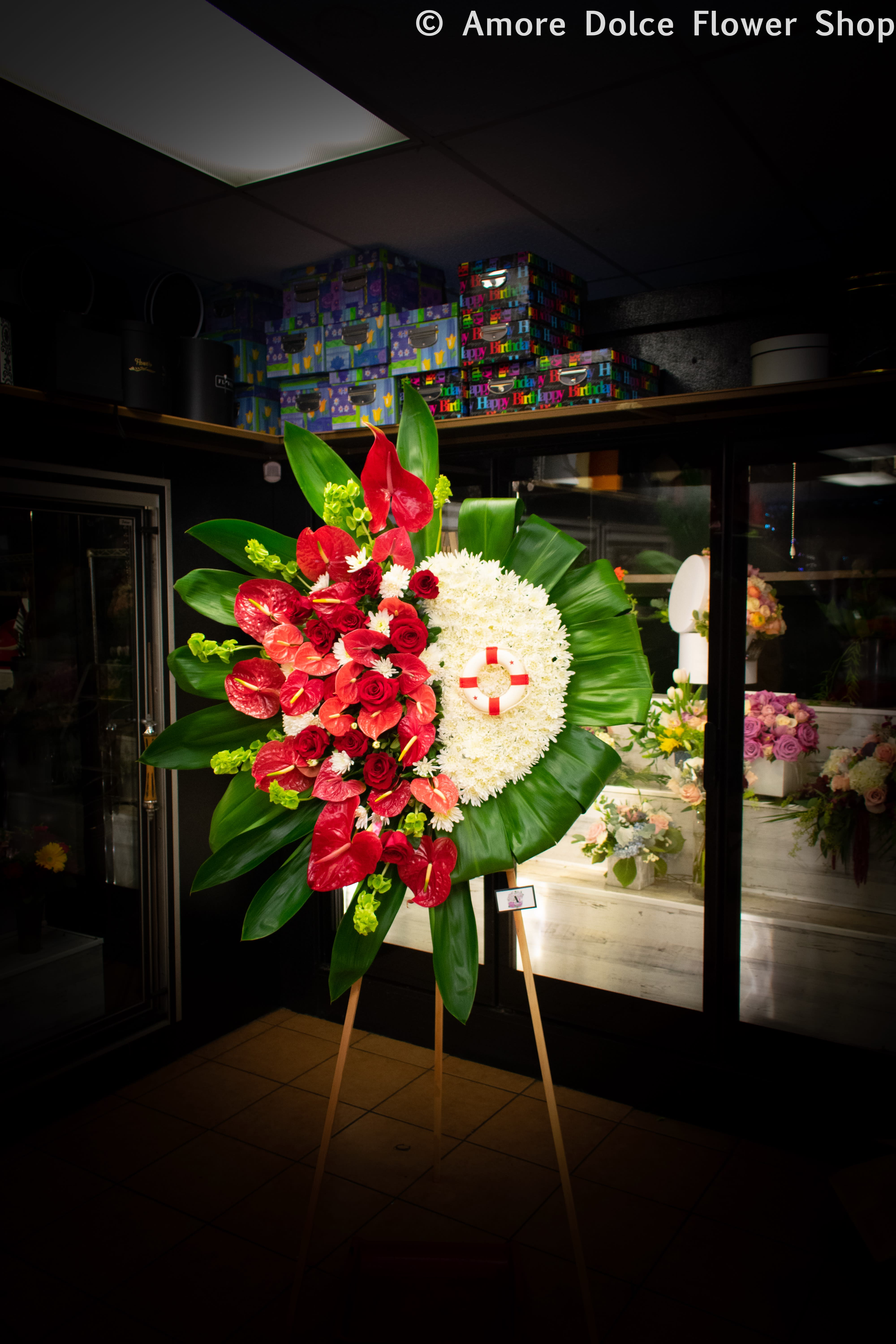 Rest in Paradise - Tropical red anthurium and long stem red rose spray on one side and white Pom Poms on the other side surrounded by tropical Ti leaves. Please note that the life preserver prop does not come included but is there to show you that we can add custom pieces to represent a little something personal about the deceased. Also please note that if you would like a sash let us know what you would like written on it in the “special instructions”section. Thank you.  