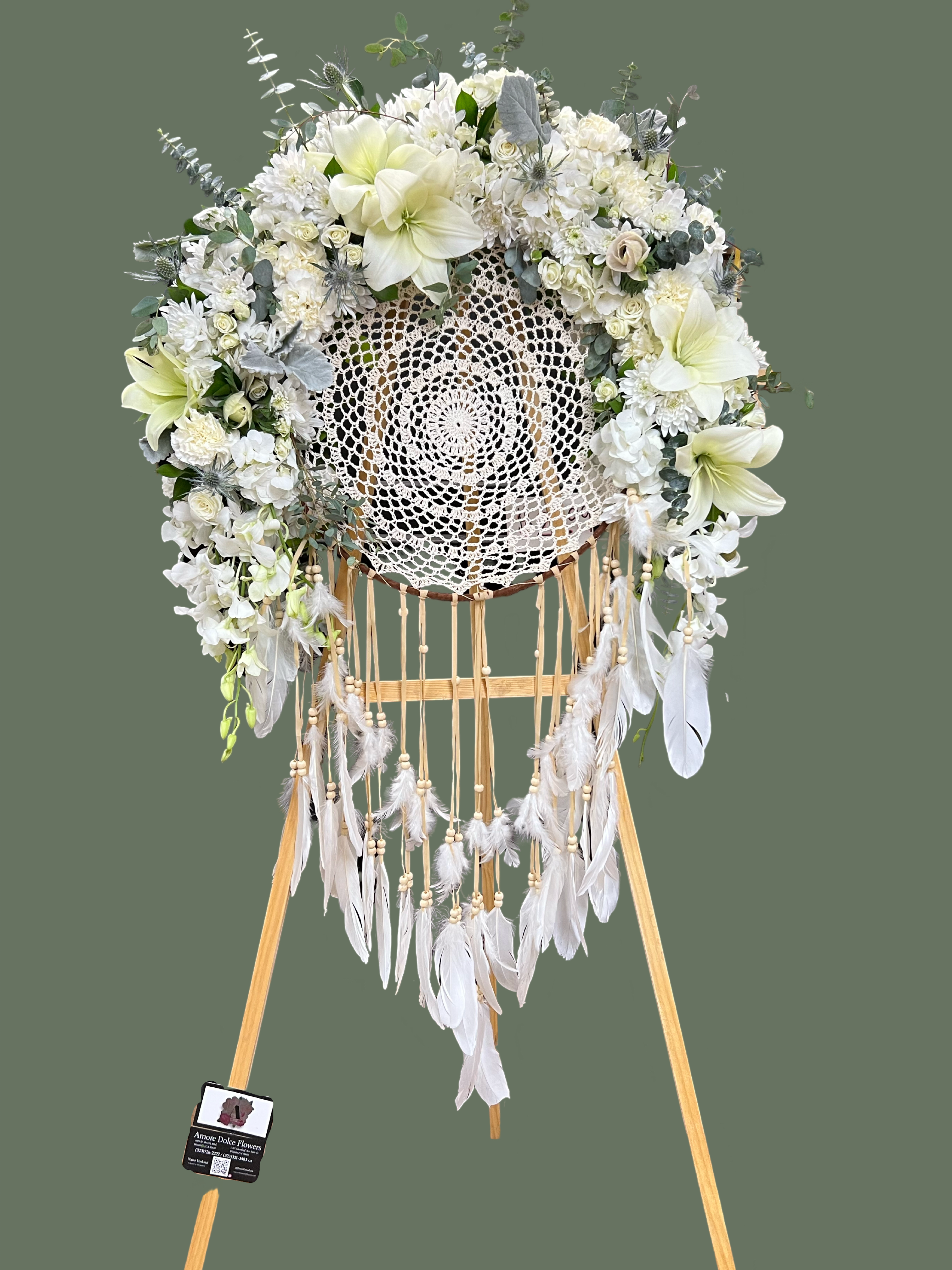 Elegant Dreamcatcher Funeral Standing Spray  - Dreamcatcher Funeral Standing Spray by Amore Dolce Flowers At Amore Dolce Flowers, we blend artistry with meaning to create one-of-a-kind floral tributes. Our Dreamcatcher Funeral Standing Spray is a stunning homage to love, peace, and remembrance. Inspired by the symbolism of dreamcatchers, this arrangement conveys comfort and serenity during moments of grief.  Featuring lush white blooms such as hydrangeas, roses, and lilies intertwined with eucalyptus and delicate foliage, the design radiates purity and elegance. The handcrafted macramé dreamcatcher centerpiece adds a unique, heartfelt touch, symbolizing the eternal weaving of cherished memories. Soft feathers and beads cascade gracefully, adding movement and depth to the design.  Perfect for funerals, memorials, or celebrations of life, this standing spray offers a deeply personal way to honor your loved one while providing comfort to family and friends.  Why Choose Amore Dolce Flowers? 	•	Unique and meaningful designs tailored to your needs 	•	Premium, fresh blooms sourced with care 	•	A dedication to craftsmanship and heartfelt service  Let us help you celebrate a life beautifully lived. Visit our store or contact us to customize this exquisite tribute for your loved one.   The Dreamcatcher is included in the price 