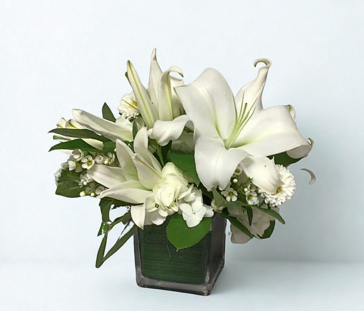 White lilies arranged in a square glass vase with small white accent flowers