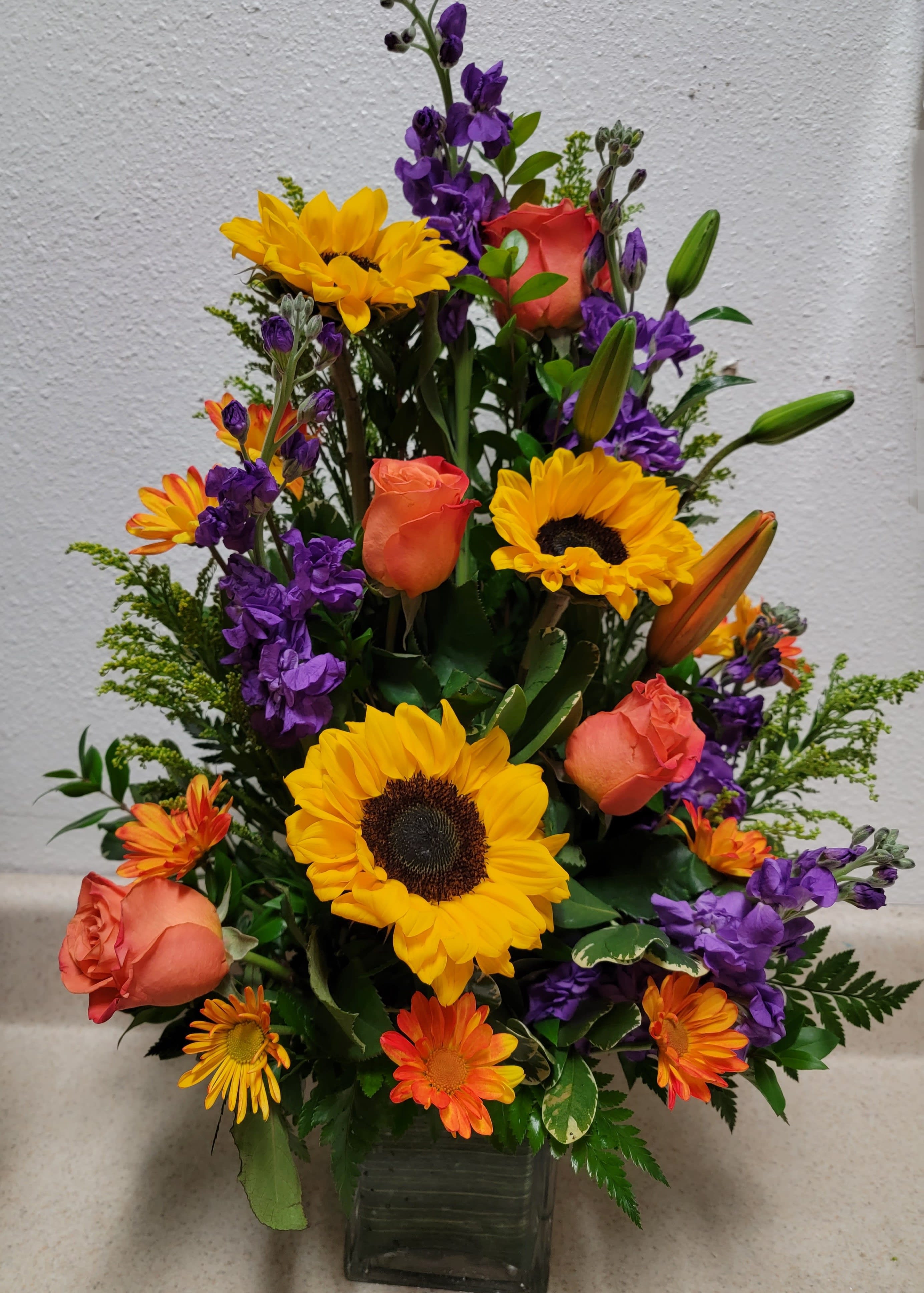 Mixed bouquet of sunflowers, orange roses, and purple flowers in a glass vase