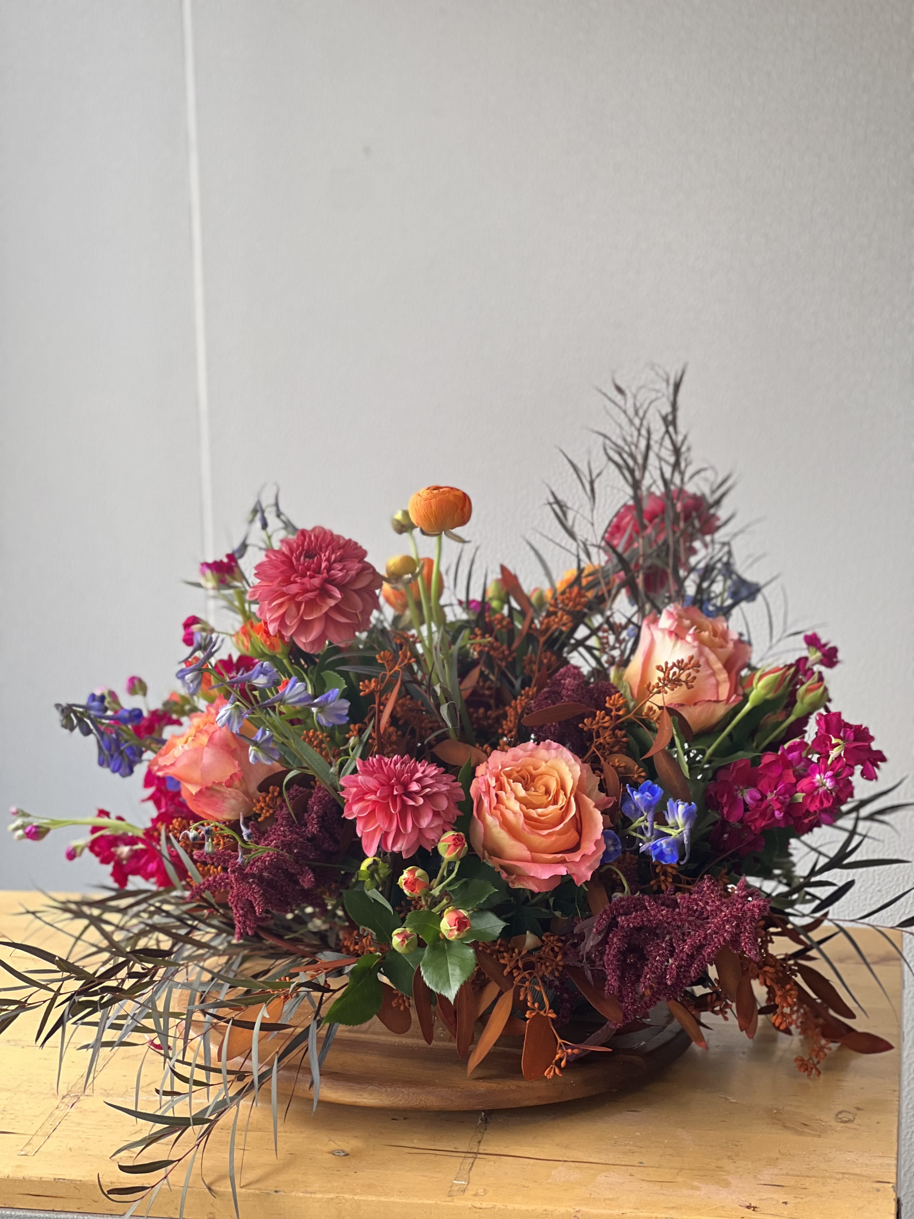 Seasonal Centerpiece  - Flowers: free spirit roses, burgundy amaranthus, royal blue delphinium, orange spray roses, orange seeded eucalyptus, mauve dahlias, magenta stock, burgundy willow eucalyptus  Arranged in a clear dish that sits gently on a table 