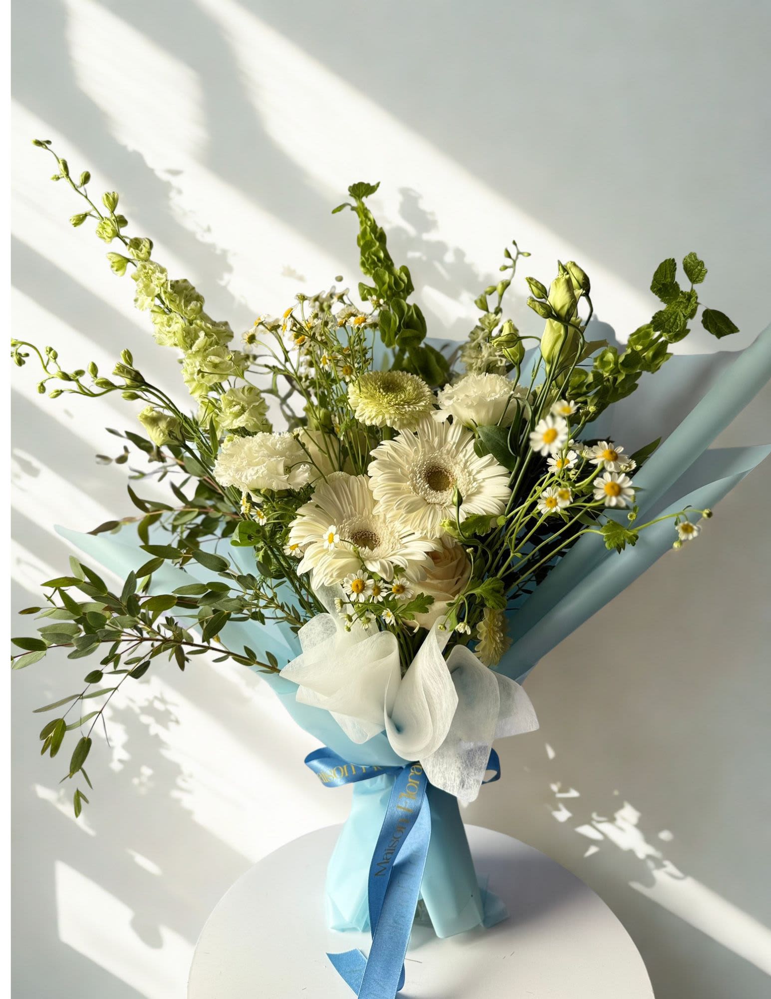 Whispering Meadow - A fresh and airy bouquet inspired by the softness of a spring morning. Whispering Meadow features elegant white gerberas, delicate chamomile daisies, creamy carnations, lisianthus, and soft green snapdragons wrapped in pastel blue for a refined yet whimsical look. Designed with movement and texture in mind, this arrangement brings a sense of calm, light, and natural beauty — perfect for birthdays, get well wishes, new beginnings, or simply brightening someone’s day.
