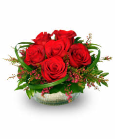 Low glass bowl of red roses with looping green blades and pink filler flowers