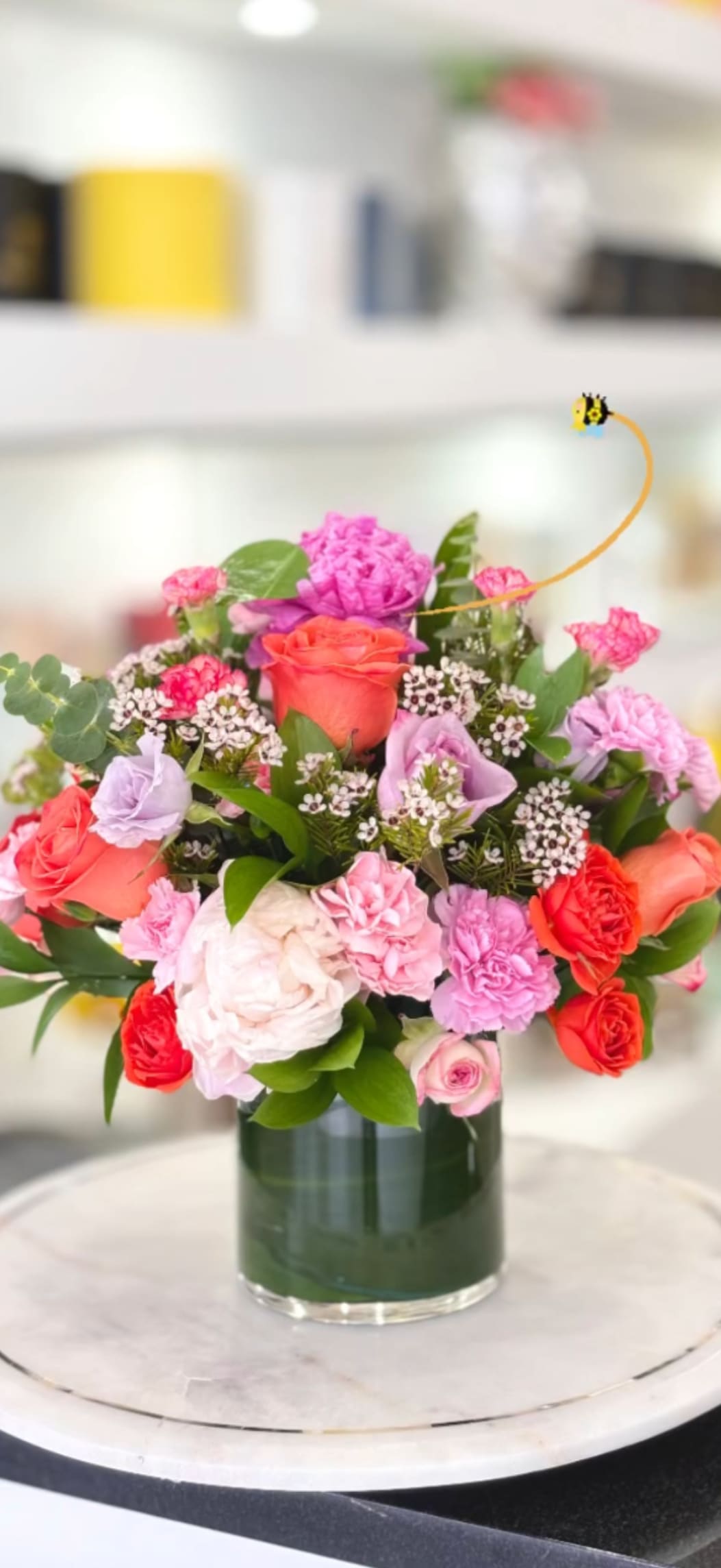 Brillante  - A lush, rounded bouquet of mixed roses and ruffled carnations that reads joyful and romantic - perfect as a birthday bouquet, anniversary flowers, or a general celebratory gift. The bright palette of pink, coral, red, orange, white and a touch of lavender gives the arrangement an upbeat, heartfelt presence that will make the person you're thinking of feel truly celebrated.  Tightly arranged in a clear cylindrical glass vase, the composition is dense and compact: garden-fresh roses take center stage while carnations and small white filler flowers add soft texture and fullness, finished with glossy green foliage. The vase keeps the design neat for tabletops or a desk and highlights the stems for a clean, modern look.  Give this to someone who enjoys vibrant, romantic color - it's the kind of arrangement that brightens a kitchen counter, office, or bedside table and reads as both intimate and festive. Care is simple: keep water fresh and recut stems every few days to prolong vase life and keep the blooms looking their best.
