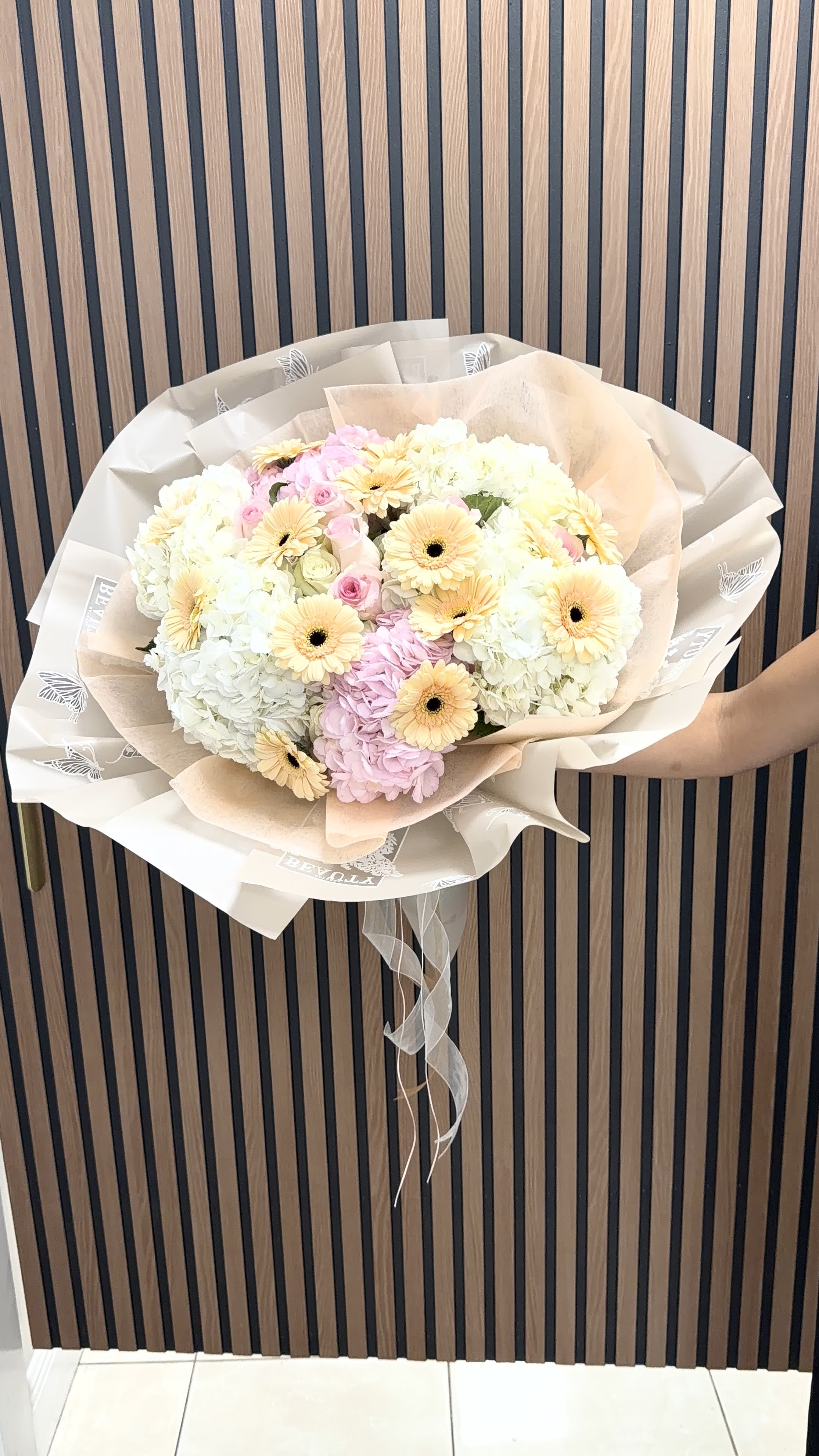 Gerberas Isle  - Soft, romantic pastels take center stage in this gerbera-and-hydrangea hand-tied bouquet-perfect as a birthday bouquet, Mother's Day arrangement, or a thoughtful anniversary gift. Gerbera daisies in pale yellow and peach mingle with full white and blush hydrangea clusters and delicate pink/blush roses to create an airy, gently rounded silhouette that reads as both celebratory and tender. The look is made for the person you want to make feel seen and cherished.  Tightly gathered and wrapped in layered cream, beige, and translucent decorative paper, the arrangement is finished with a sheer ribbon tail for a polished, gift-ready presentation. Textures contrast beautifully: the plush, cloud-like hydrangeas, the smooth petals of roses, and the cheerful faces of gerberas combine for visual depth and a soft fragrance. Sized as a generous hand-tied bouquet, it's an easy-to-hold statement that arrives ready to display or pass on.  Lovely for birthdays, thank-you gestures, spring celebrations, and anniversary moments, this bouquet reads as both everyday sweetness and special-occasion beauty-she'll feel genuinely celebrated. For best enjoyment, keep stems in fresh water, away from direct heat or draft, and refresh the water every couple of days to extend vase life.