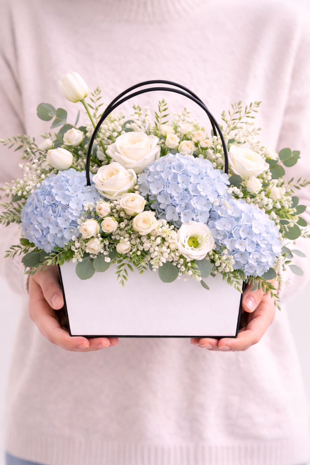 Coastal Breeze  - an airy garden- style arrangement featuring soft blue hydrangeas, creamy white roses, delicate seasonal blooms, and fresh greenery in our signature floral tote. Elegant, peaceful, and effortlessly timeless. 